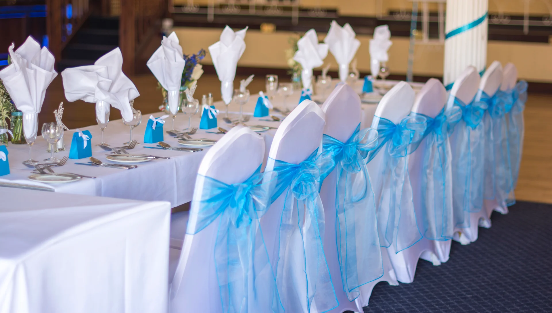 An elegantly set top table featuring white linens, matching chair covers adorned with vibrant blue organza sashes, and neatly arranged place settings.