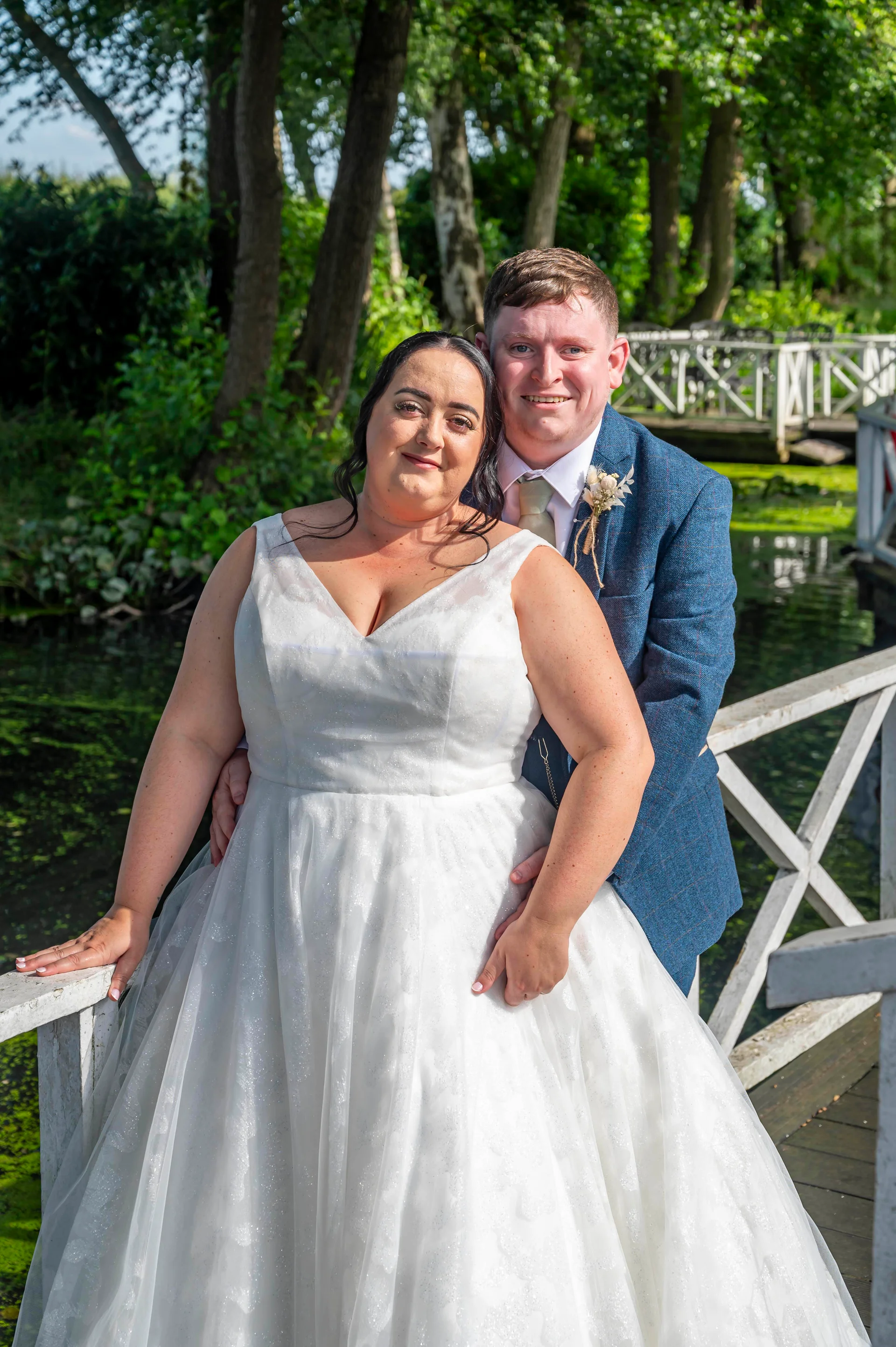 A smiling bride in a sparkly white a line gown and the groom in a blue tweed suit pose together on a white wooden bridge by a lake.
