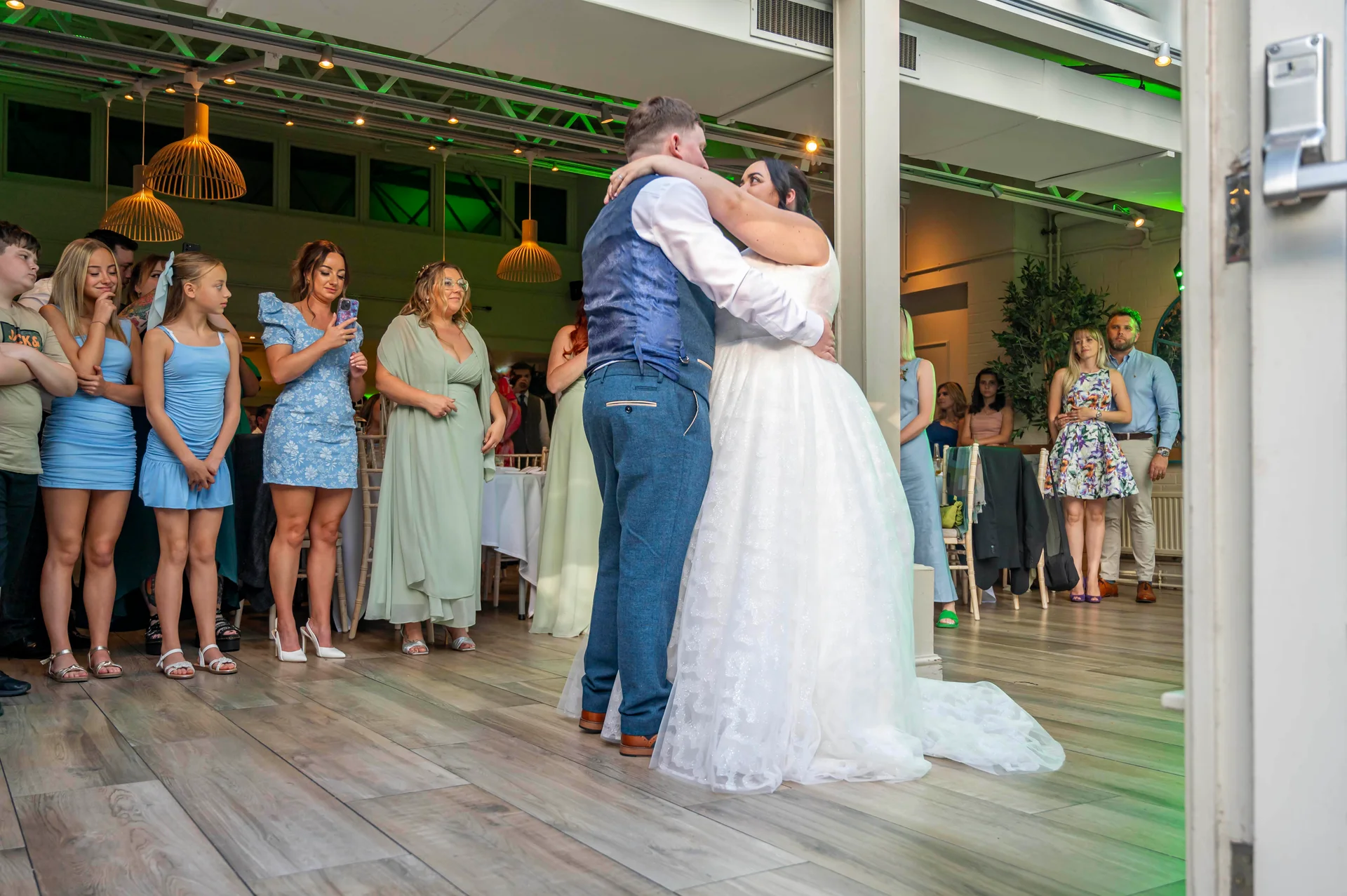 The newly married couple share their first dance inside a modern venue while guests gather around to watch.