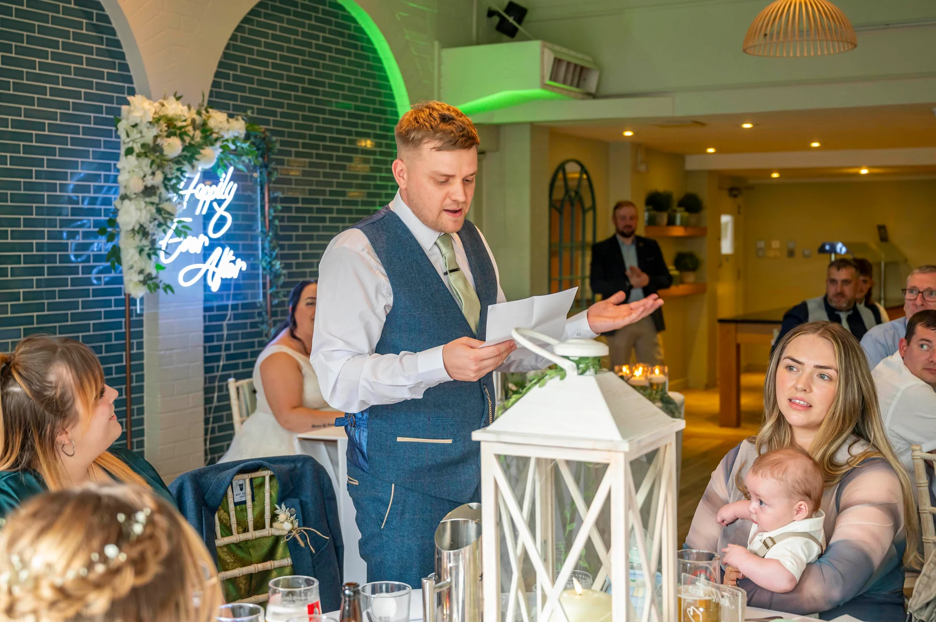 A groom delivers a speech during the wedding reception while holding a paper, with guests and a baby seated around the candlelit tables.