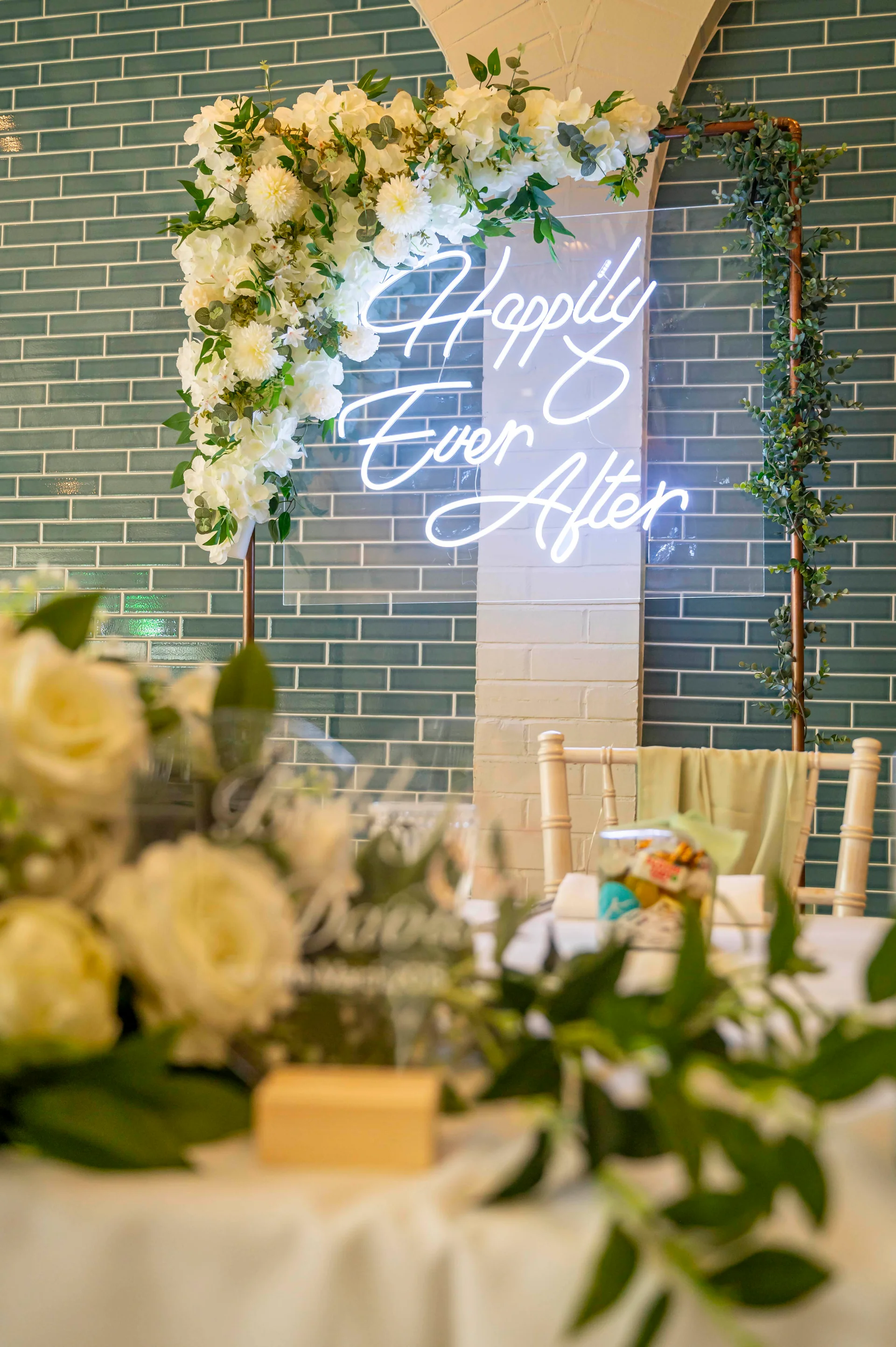 A wedding reception top table features a glowing neon sign reading Happily Ever After mounted on a copper frame and adorned with white florals and greenery.