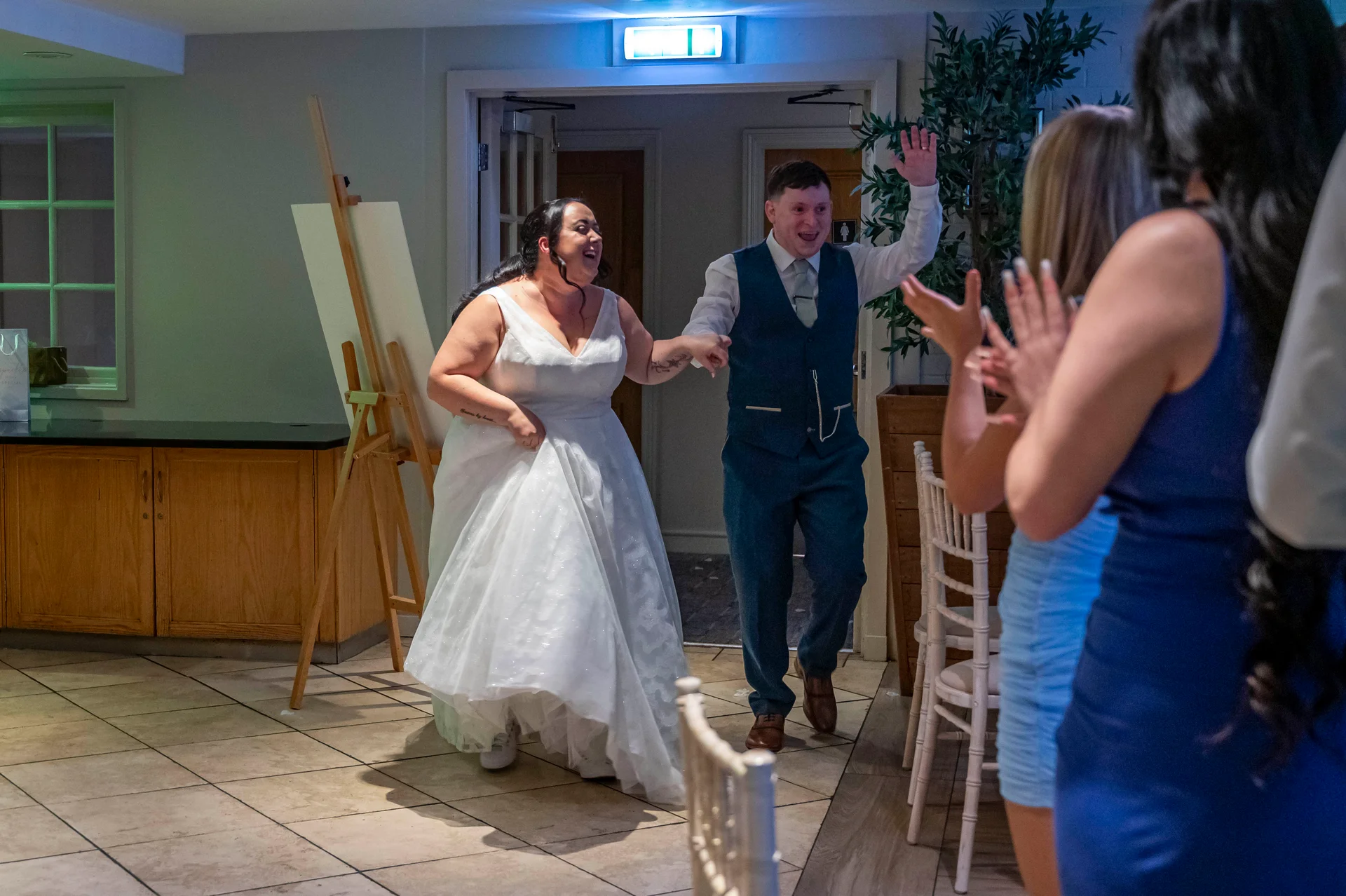 A joyful newlywed couple enter the reception holding hands while cheering wedding guests applaud them.