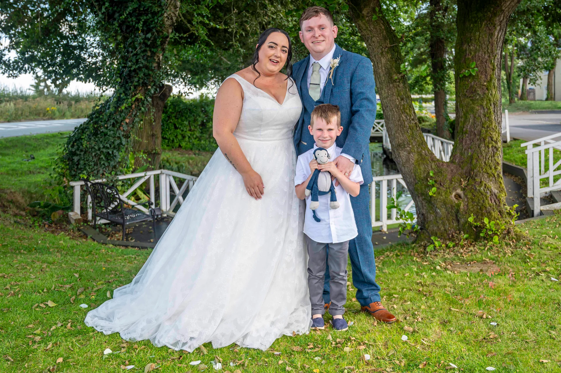 A smiling bride in an A-line wedding dress, groom in a blue tweed suit, and young page boy holding a toy monkey pose together outdoors on a lush green lawn.
