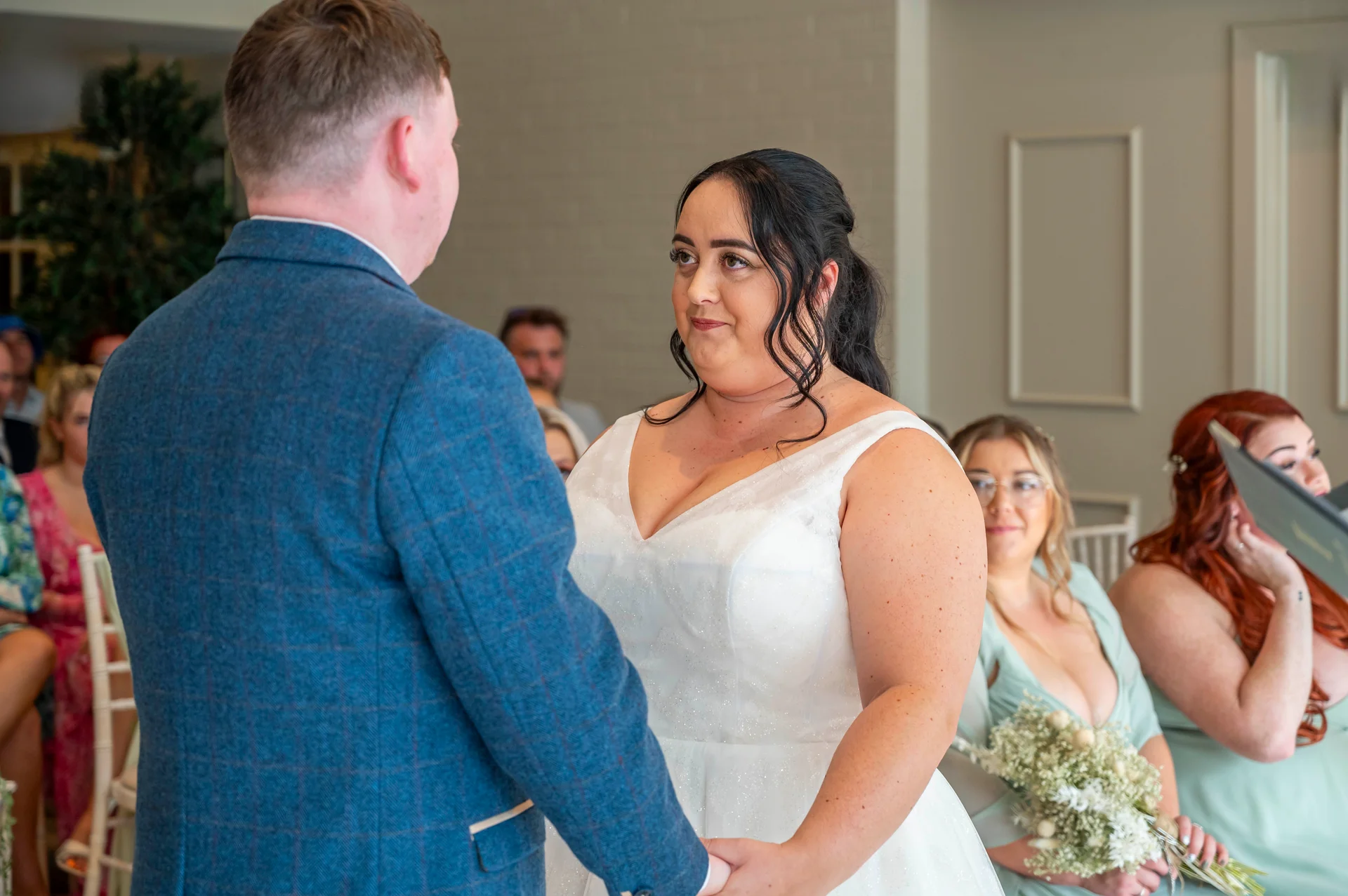 The bride looks lovingly at the groom holding her hands during their wedding ceremony while seated guests watch on.