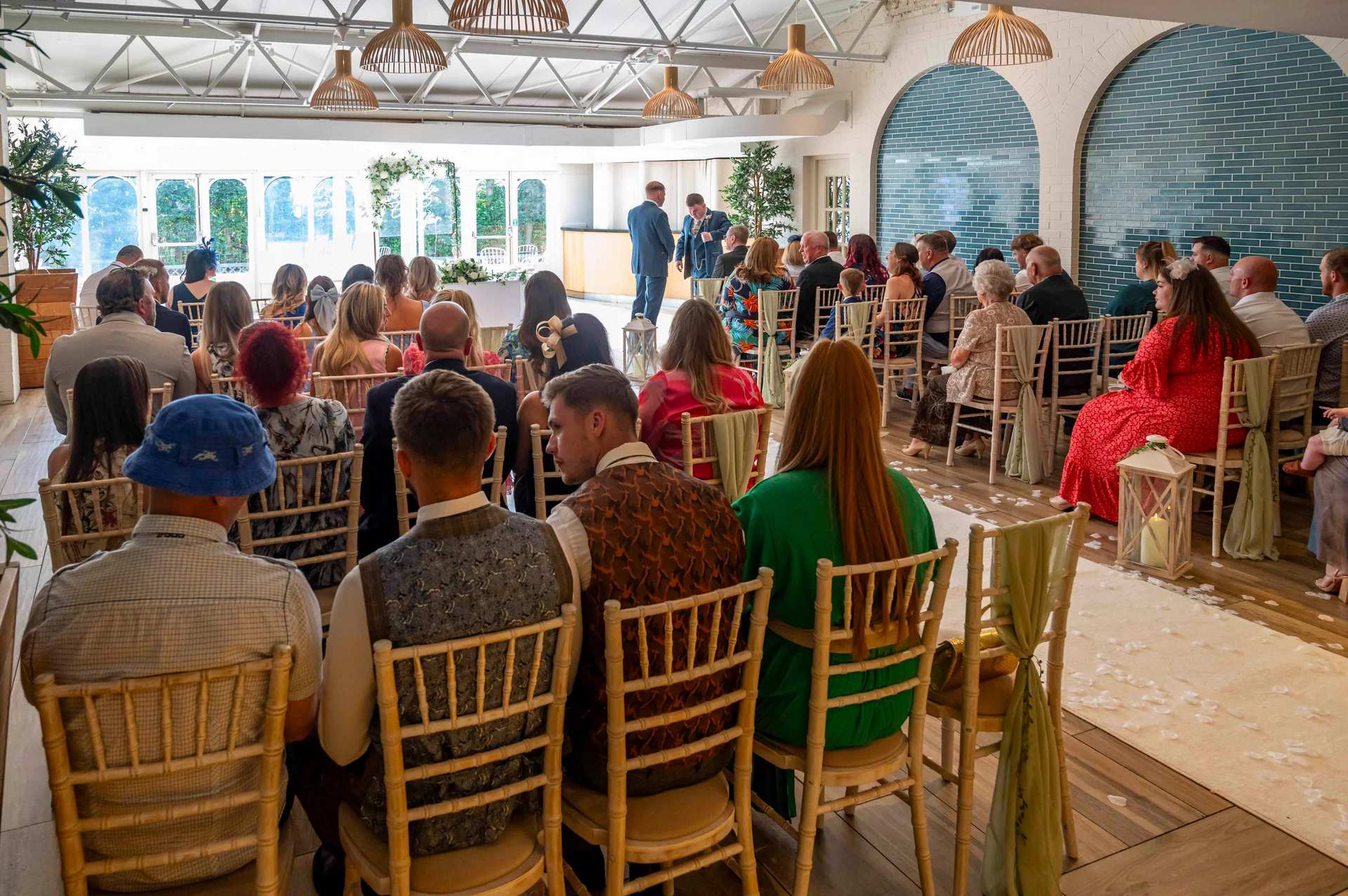 A wide shot of guests seated on chiavari chairs during an indoor wedding ceremony in a modern venue with pendant lighting and tiled arches.