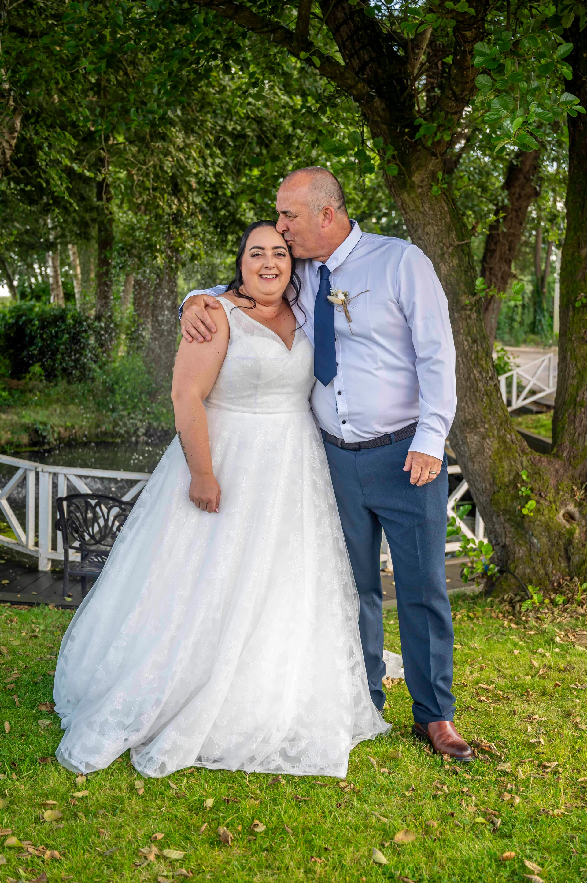 A groom gently kisses the smiling bride on the forehead outdoors in a lush garden setting beneath a leafy tree.