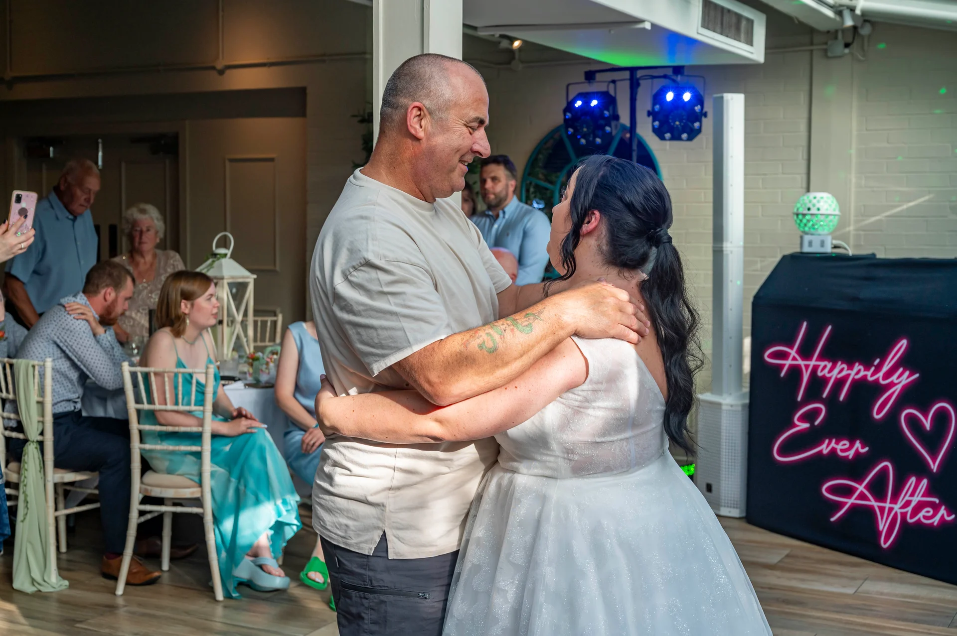 An older couple shares a joyful embrace and dance during a wedding reception, with seated guests and a glowing neon sign in the background.