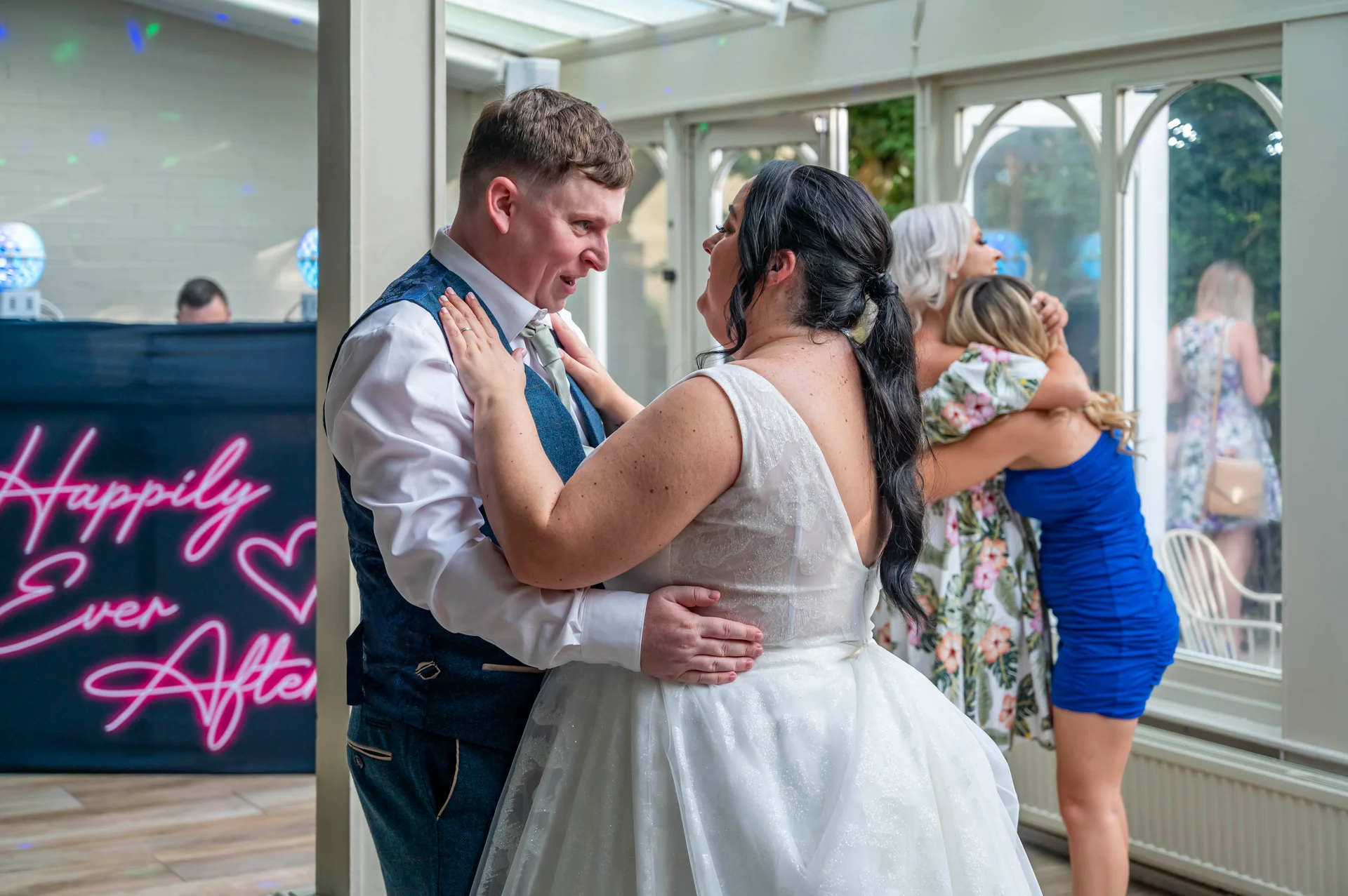 A newlywed couple laugh joyfully together during their first dance inside a bright conservatory wedding venue featuring a neon sign.
