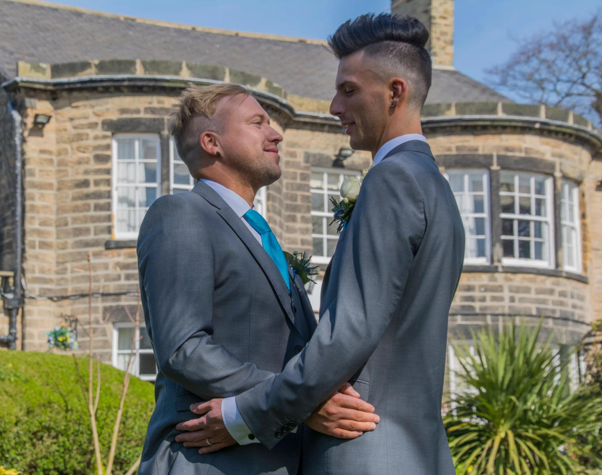 Two grooms in matching grey suits and bright blue ties lovingly embrace outside a historic stone manor house on a sunny day.
