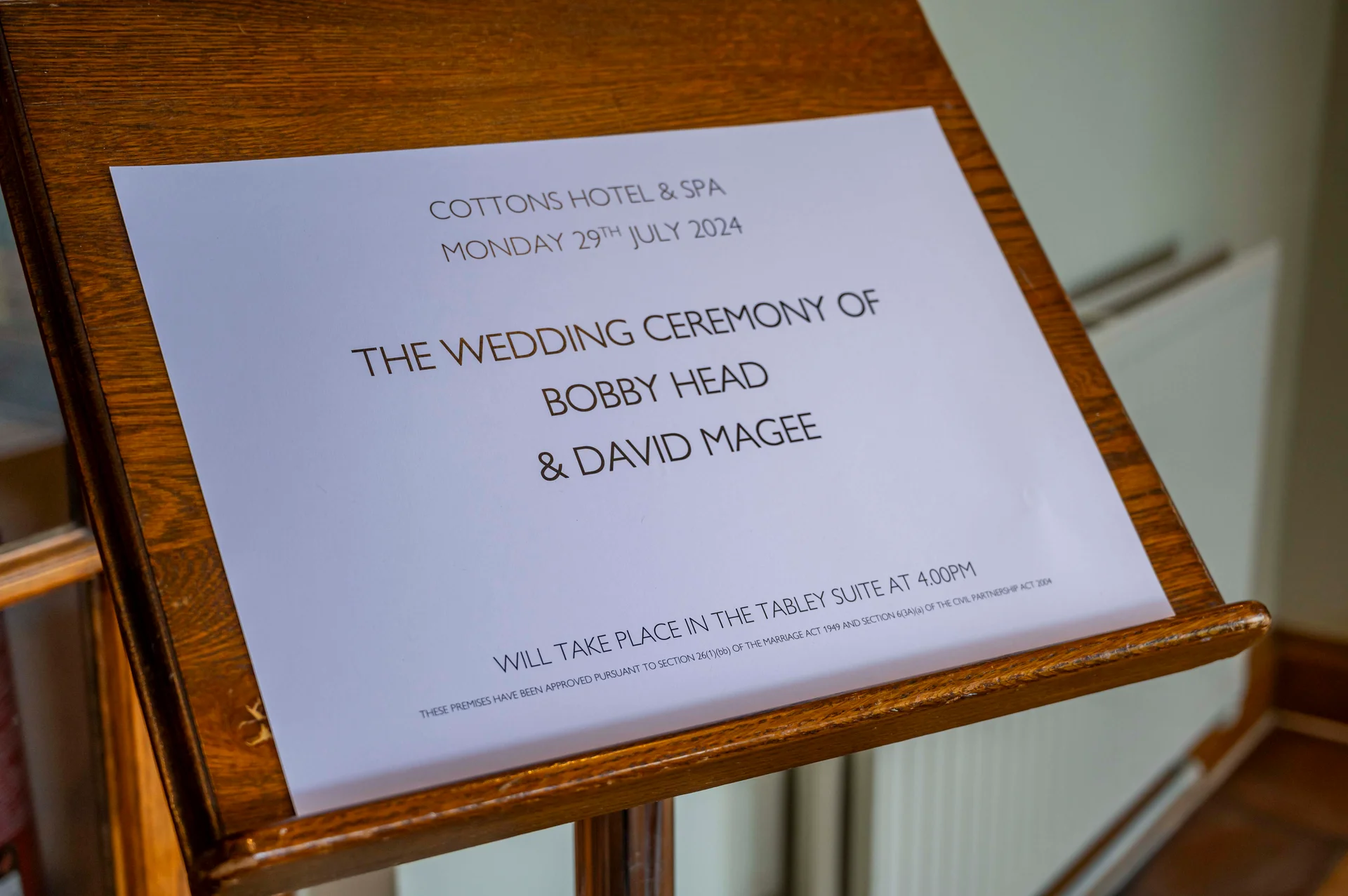 A printed wedding ceremony notice for Bobby Head and David Magee is displayed on a wooden easel stand inside a hotel venue.