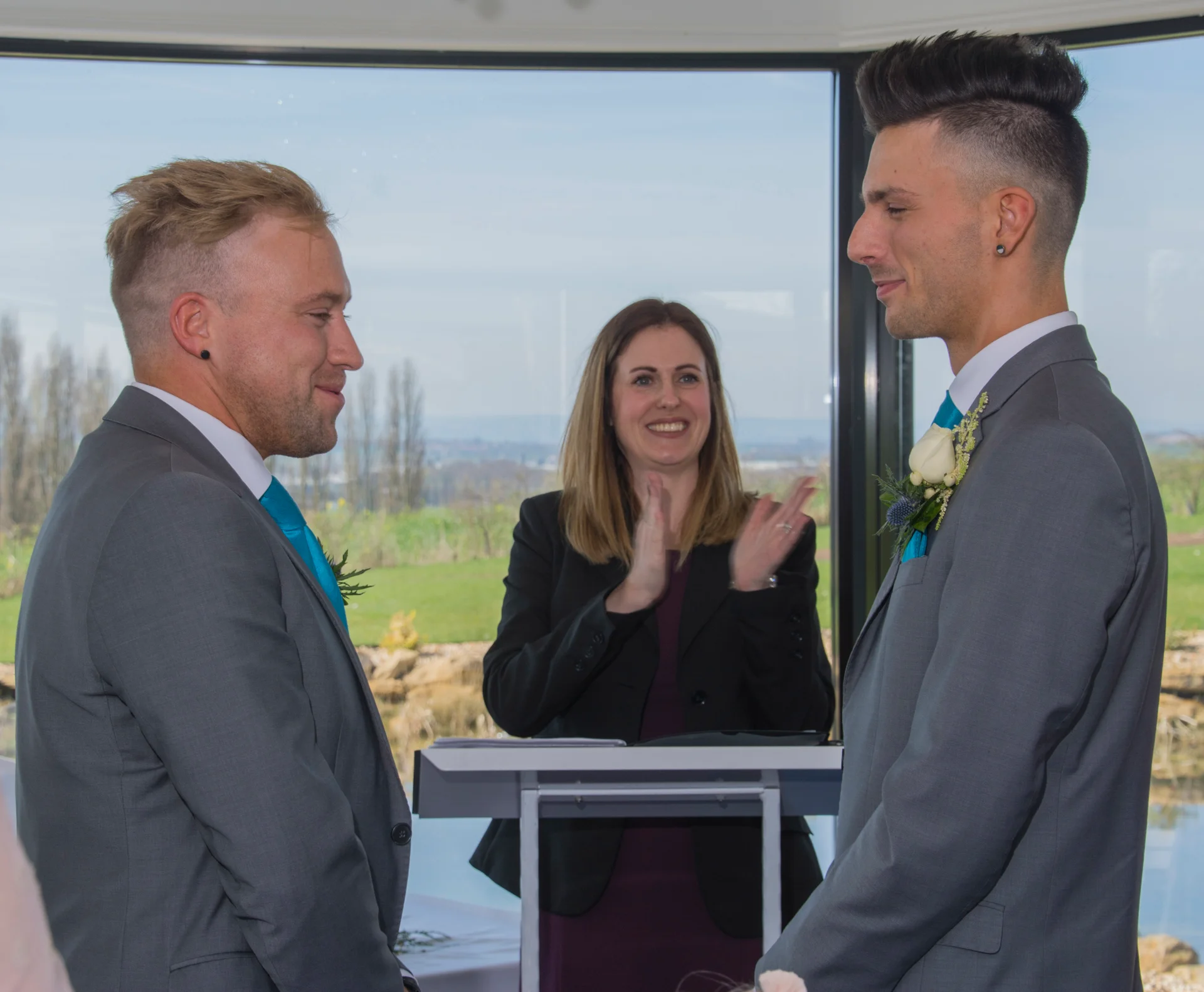 Two grooms in matching grey suits and turquoise ties stand happily at the altar during their civil ceremony as the celebrant applauds.