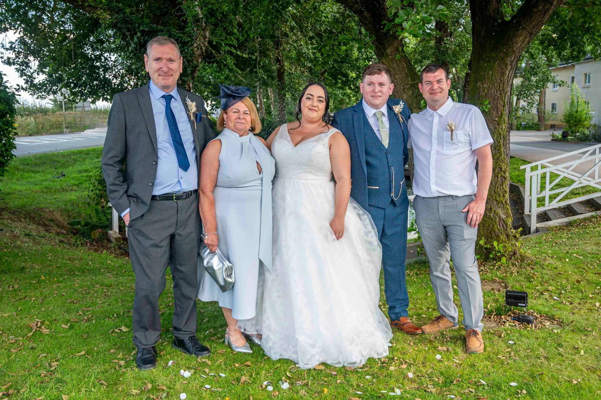 A formal family group photograph of the bride and groom posing outdoors on a lush green lawn next to trees with their family members.