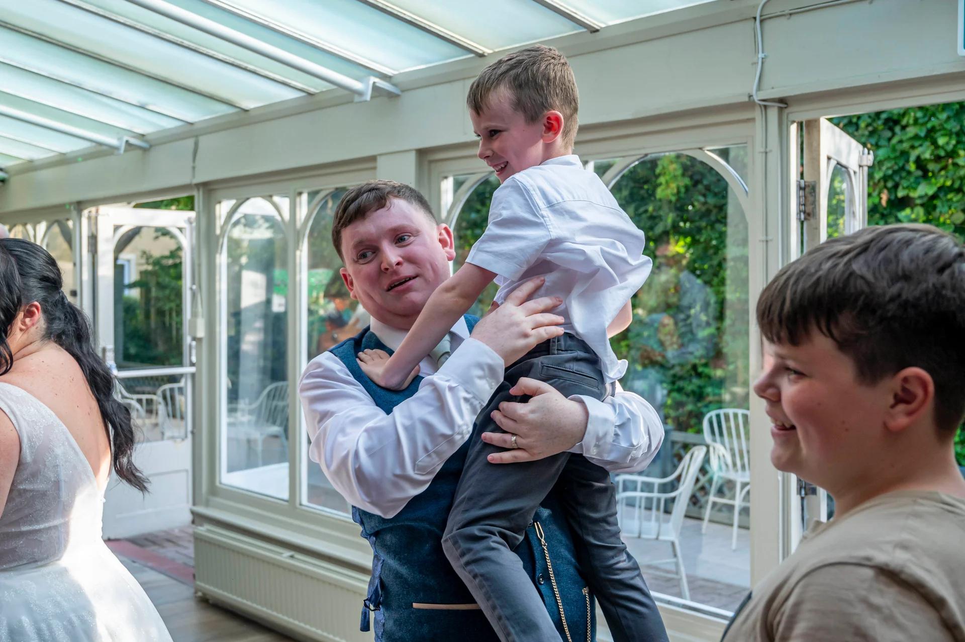 A groom lovingly lifts a smiling young boy into the air inside a bright conservatory while guests look on joyfully.