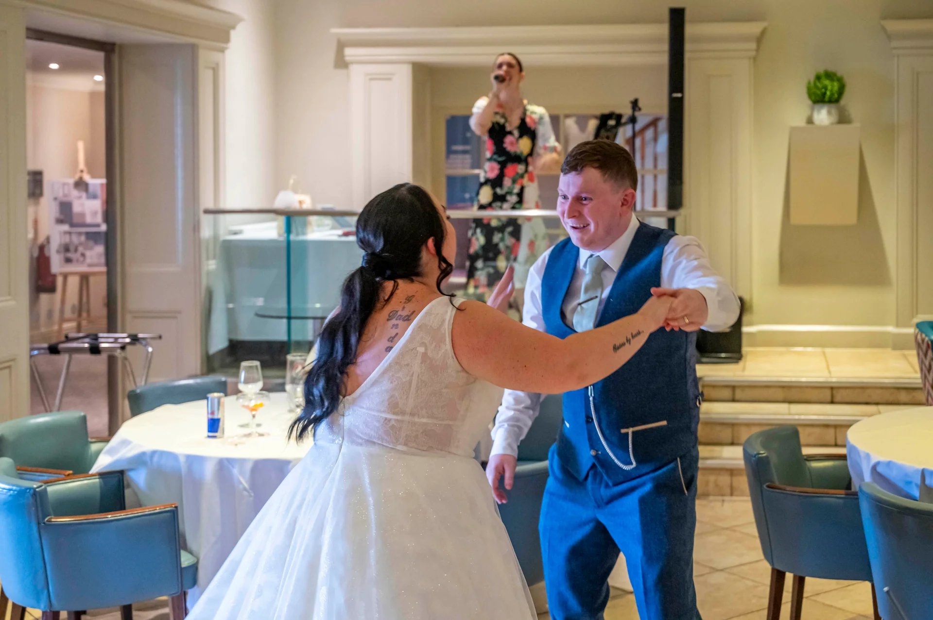 A newlywed couple shares a joyful first dance inside a brightly lit restaurant reception venue, with the bride in a glittering gown and the groom in a blue waistcoat.