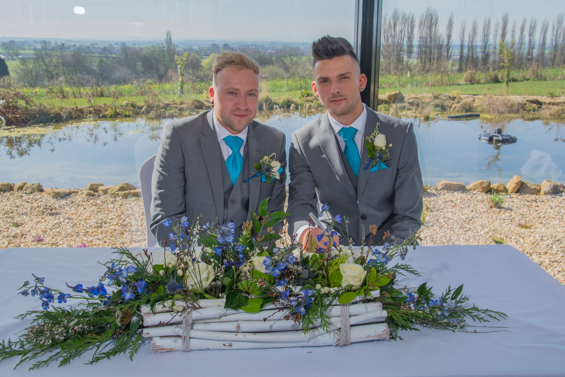Two grooms in matching grey three-piece suits smile while signing the register at a table adorned with blue and white floral arrangements against a scenic lakeside window backdrop.