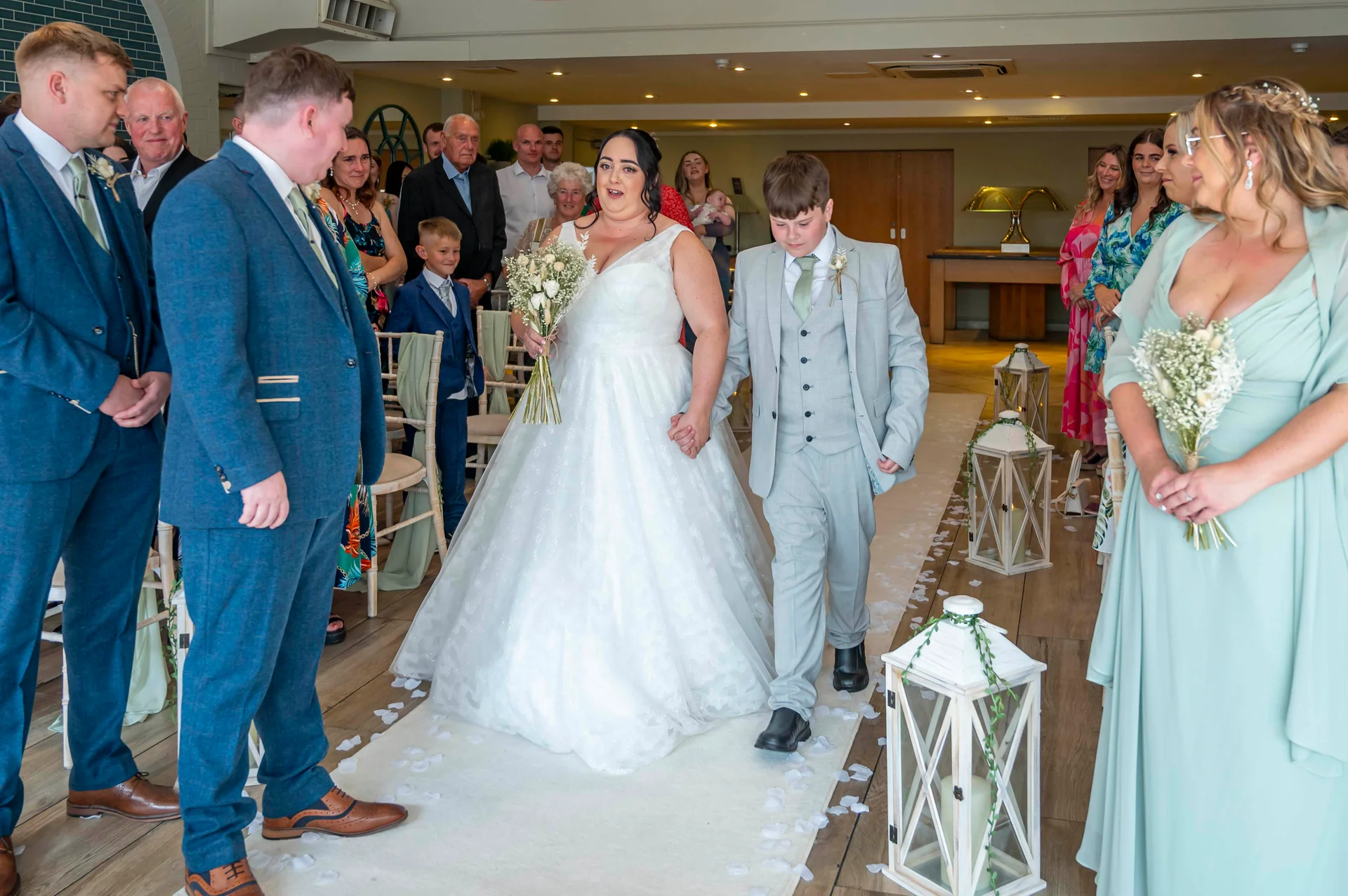 A joyful bride walking down the aisle hand in hand with a page boy toward the groom, flanked by guests and decorative lanterns.
