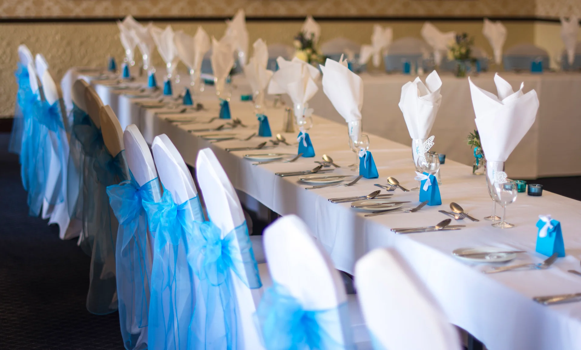 A long wedding banquet table is elegantly set with crisp white tablecloths, silverware, and vibrant turquoise sashes tied to the chair covers.