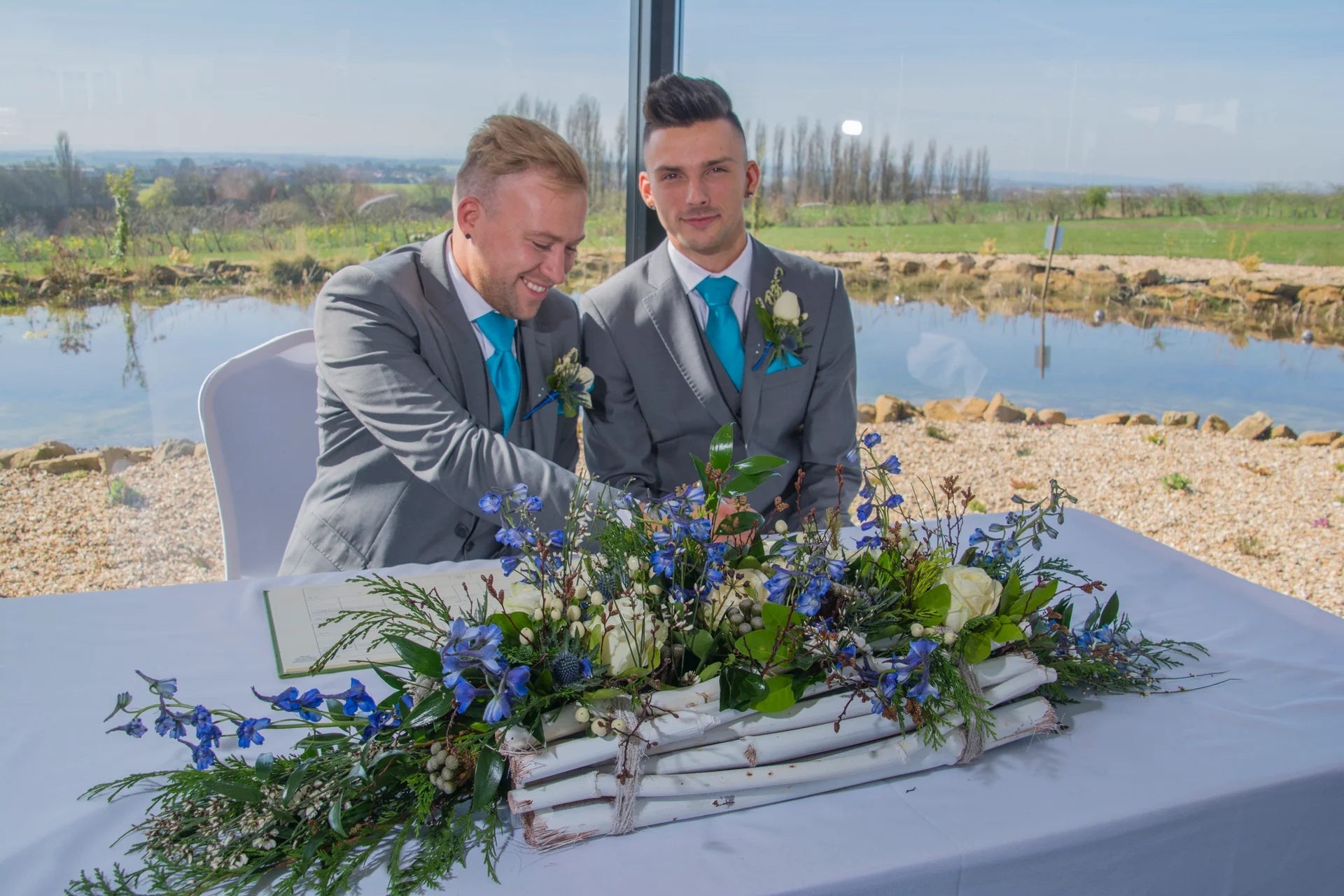 Two grooms in matching grey suits and bright blue ties sign the wedding register behind a table adorned with blue delphiniums and white roses, with a scenic lake visible through the window.