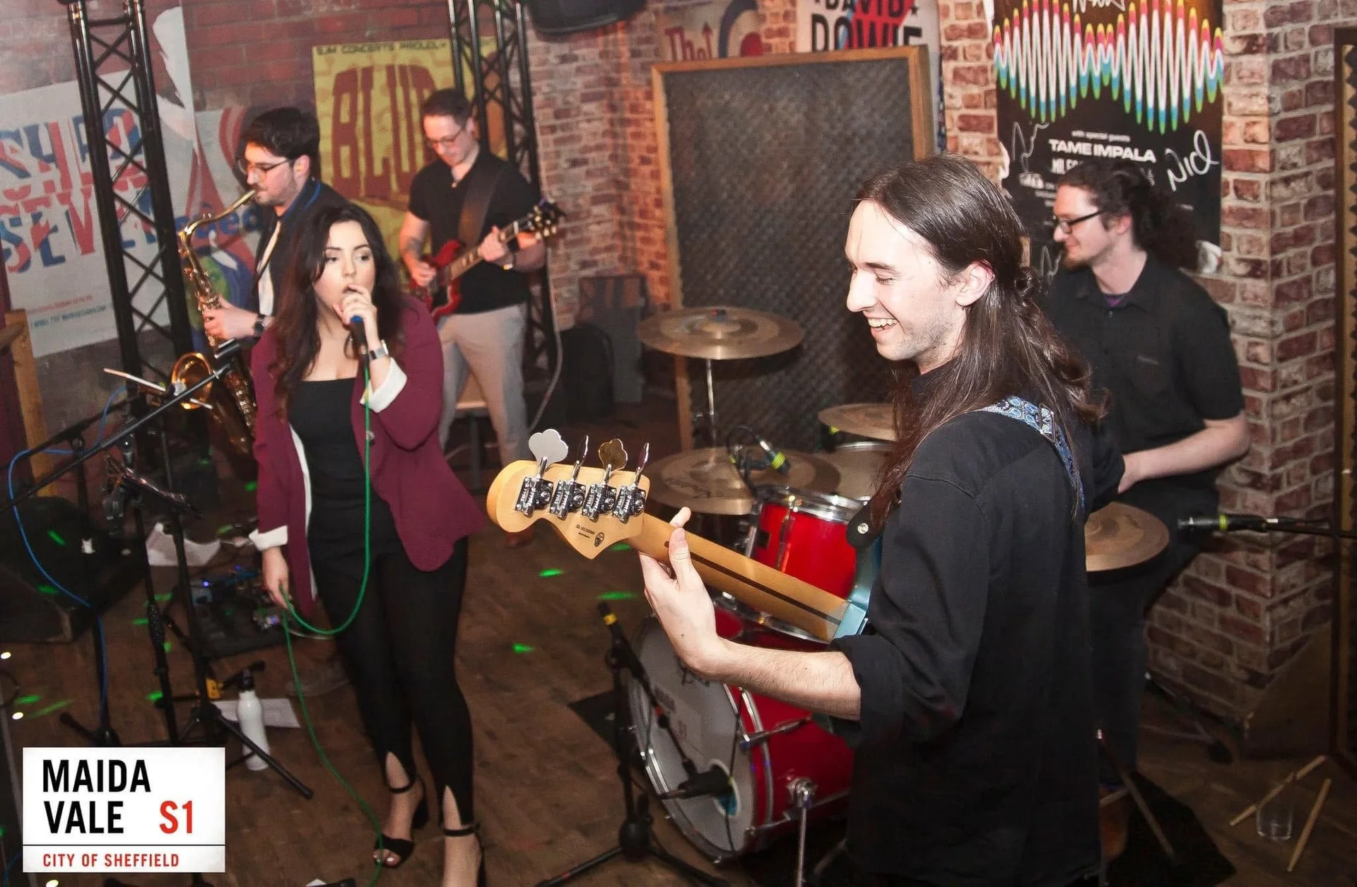 A live function band performs on stage with a female singer, a bass guitar player smiling forward, and band members playing saxophone and drums in an industrial venue with exposed brick walls.