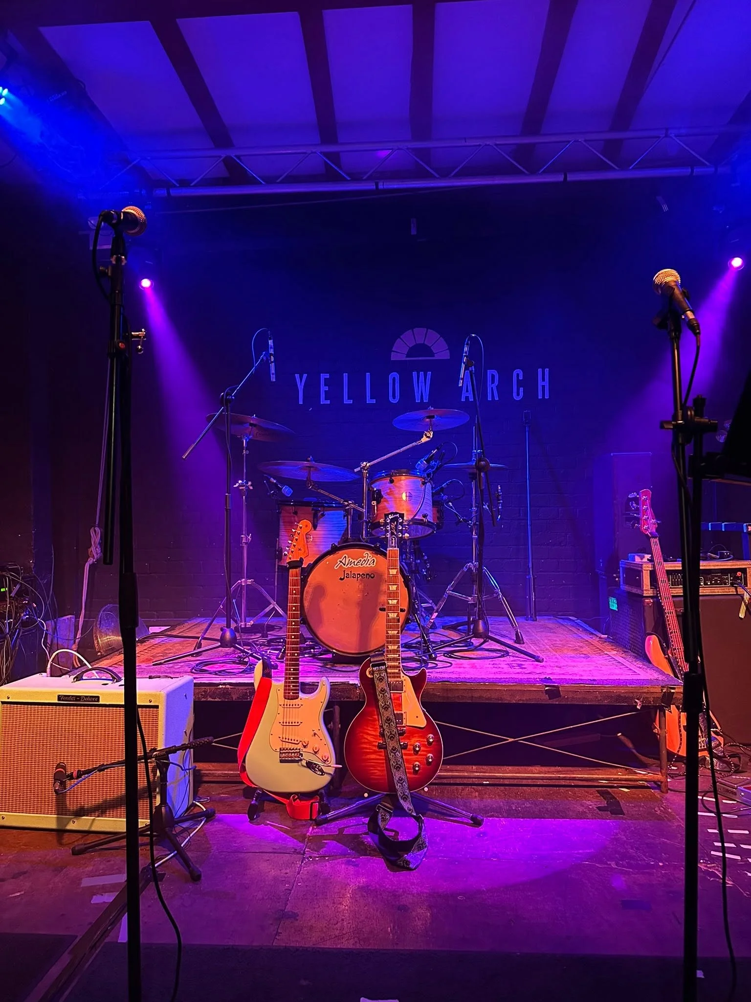 A wedding stage is set up with instruments including guitars, a drum kit, and microphones, illuminated by dramatic blue and purple stage lighting.