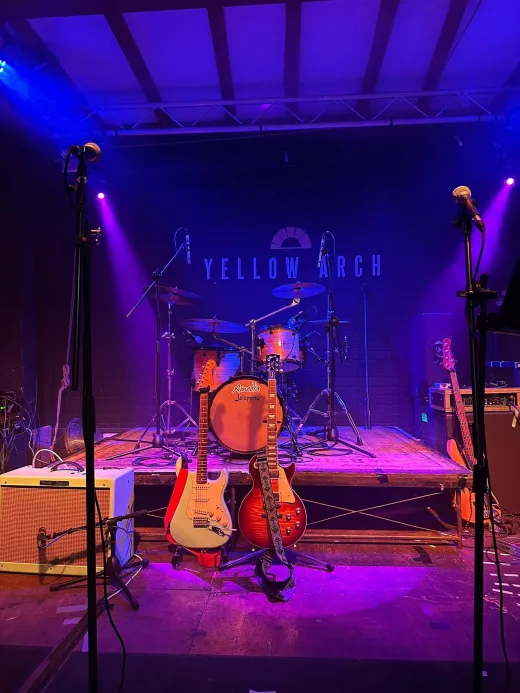 A wedding stage is set up with instruments including guitars, a drum kit, and microphones, illuminated by dramatic blue and purple stage lighting.