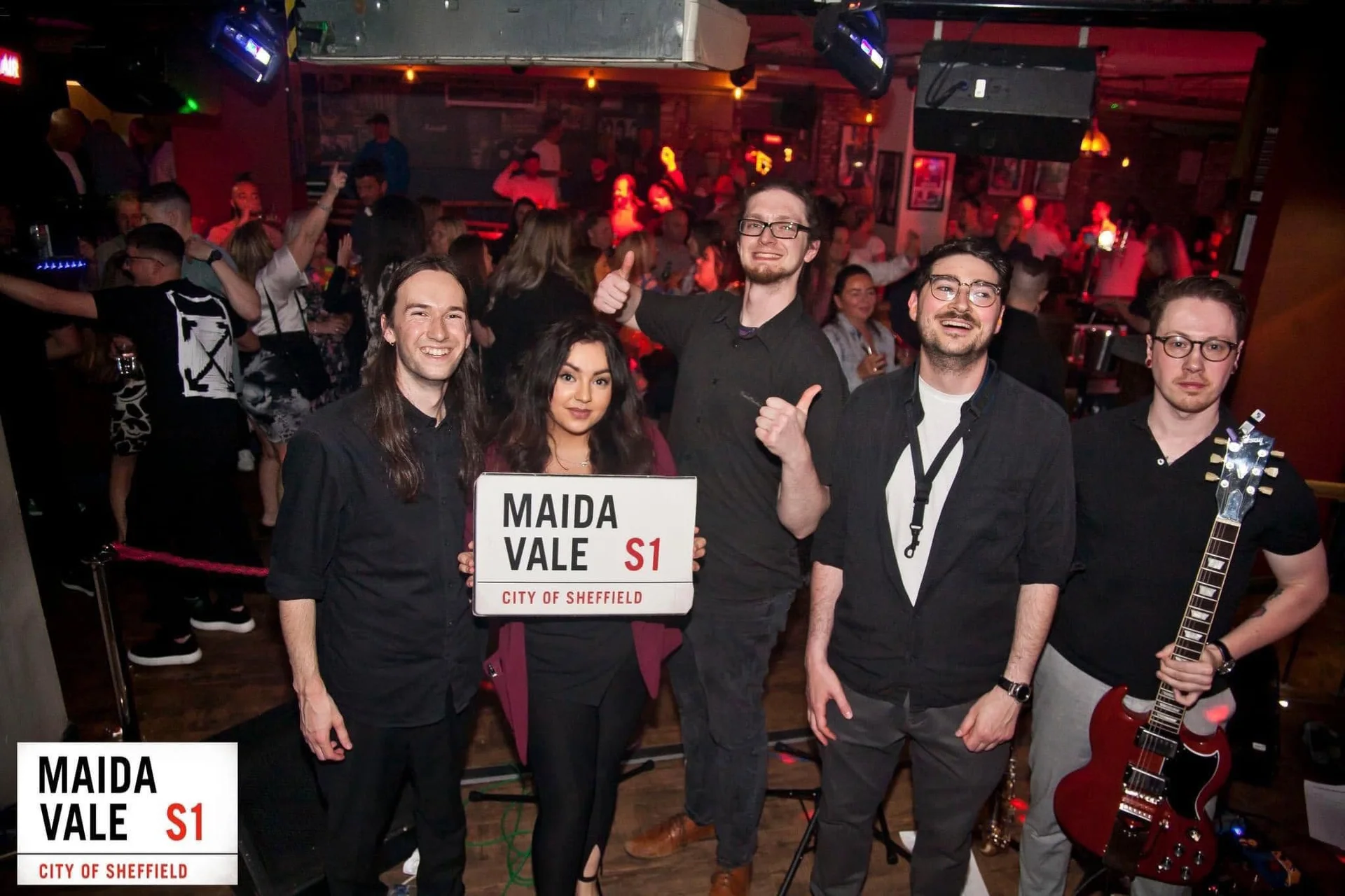 A lively wedding band poses together on stage holding a street sign in a crowded evening venue.