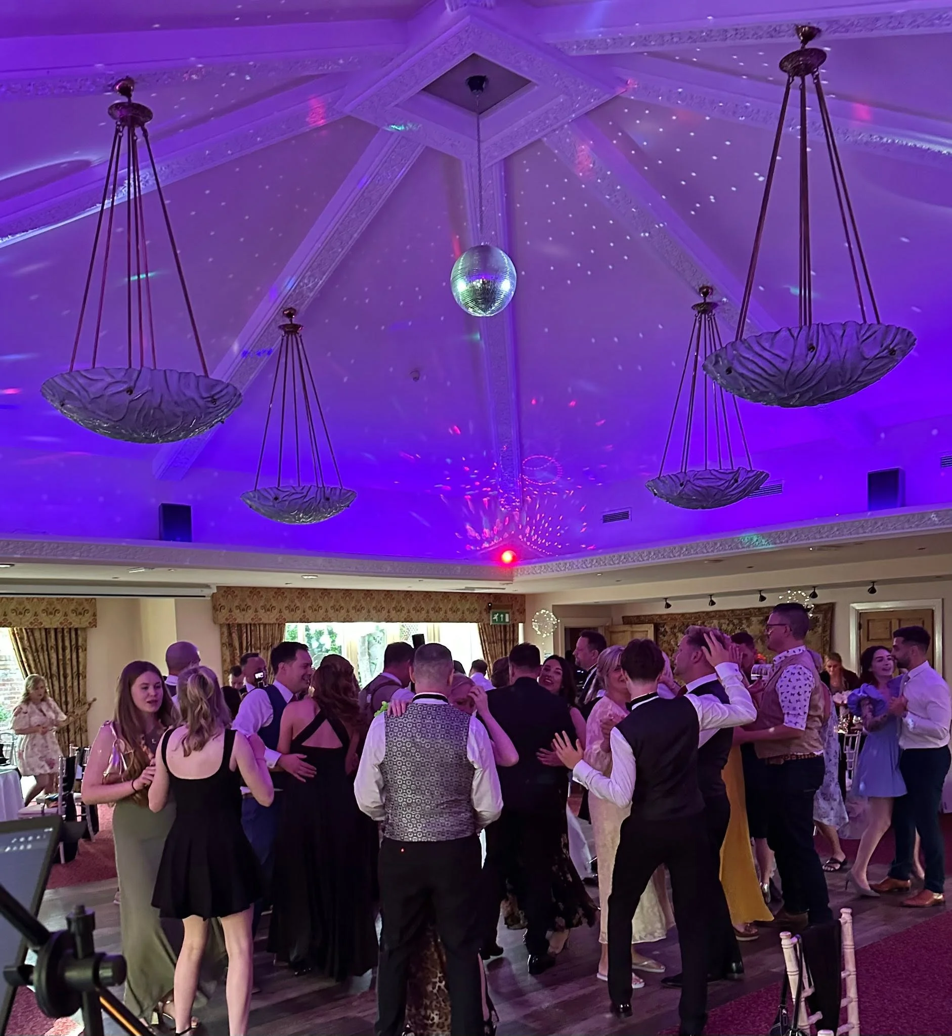 A lively wedding reception with guests dancing on a packed dance floor under a vaulted ceiling illuminated by disco lights and large pendant fixtures.