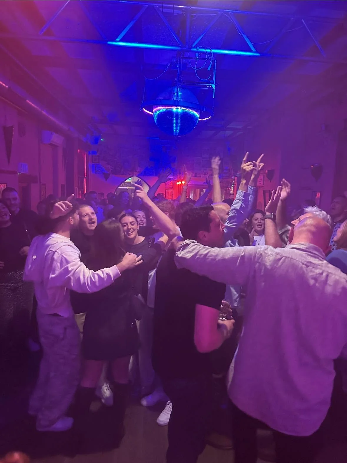 A lively wedding party dancing on a packed dance floor illuminated by vibrant blue and purple disco lights beneath a hanging disco ball.