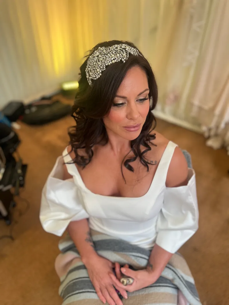 A beautiful bride sitting indoors during morning preparations, wearing an embellished crystal headband and an off-the-shoulder gown with puffy sleeves.