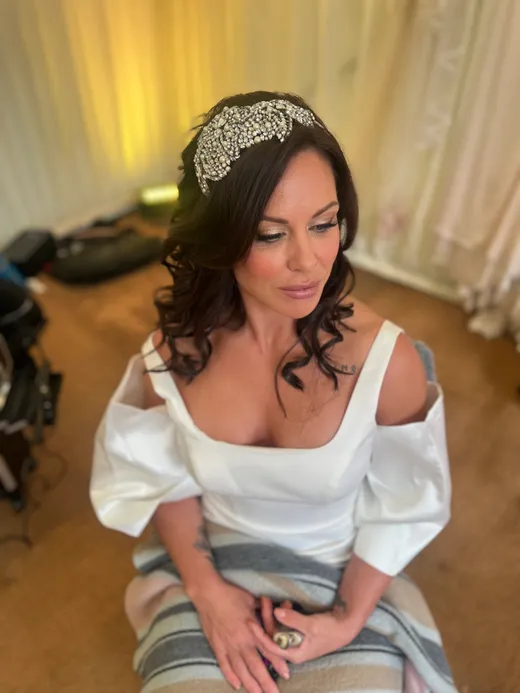 A beautiful bride sitting indoors during morning preparations, wearing an embellished crystal headband and an off-the-shoulder gown with puffy sleeves.