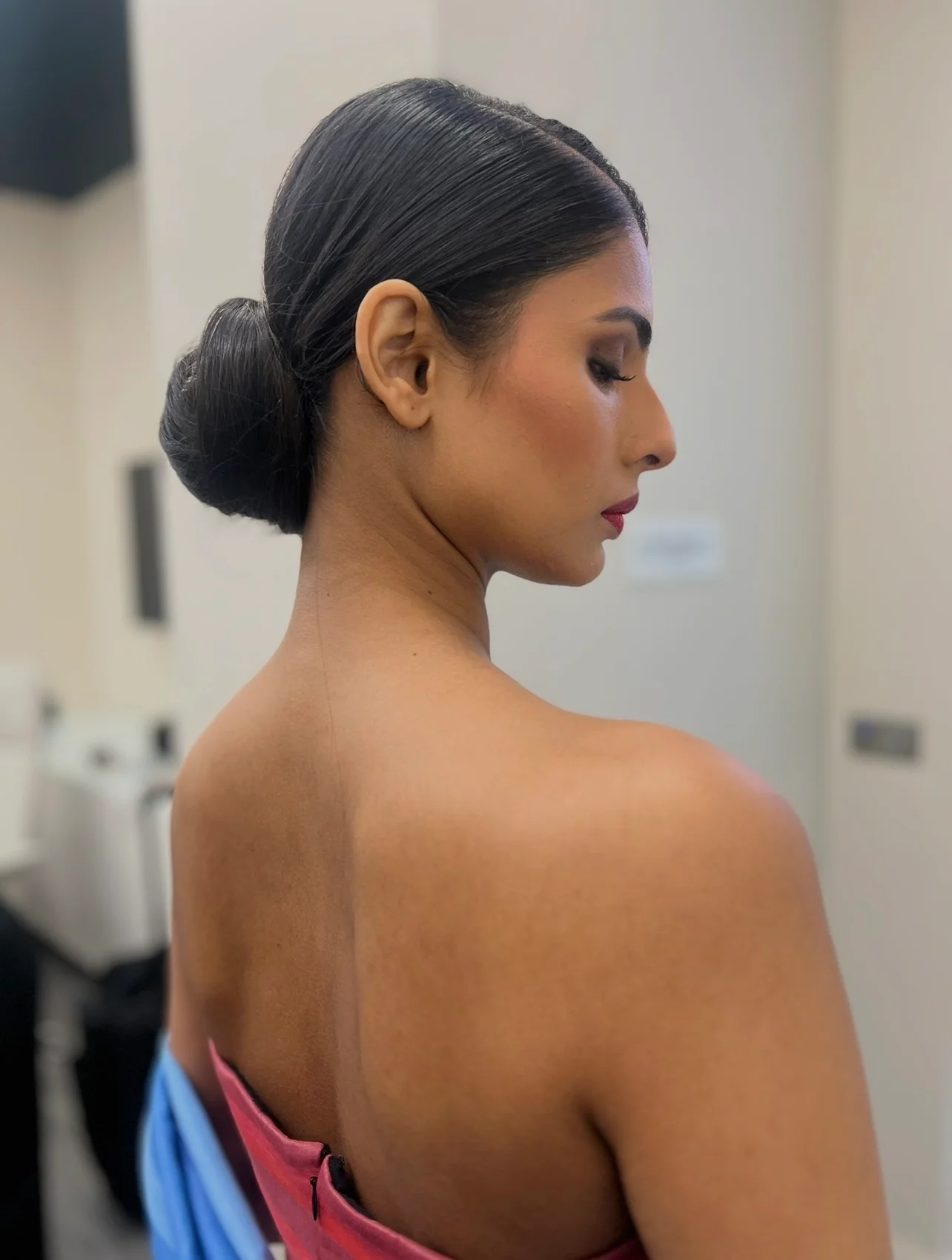 A close-up back view of a bride showcasing her elegant sleek bun hairstyle and flawless soft glam makeup with a bold red lip.