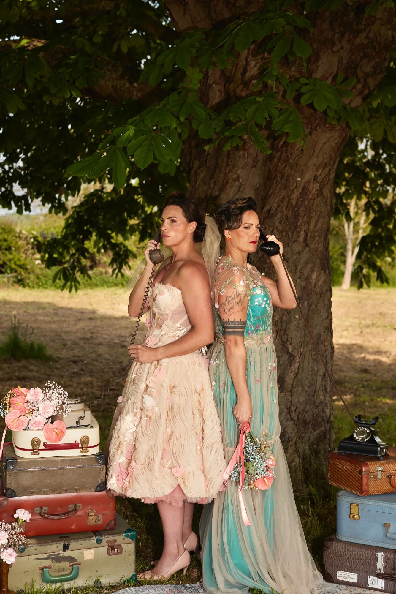 Two brides pose back to back under a large tree holding vintage telephones, surrounded by stacks of old suitcases and floral arrangements outdoors.