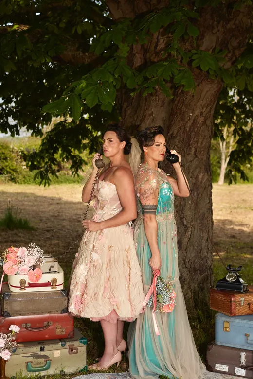 Two brides pose back to back under a large tree holding vintage telephones, surrounded by stacks of old suitcases and floral arrangements outdoors.
