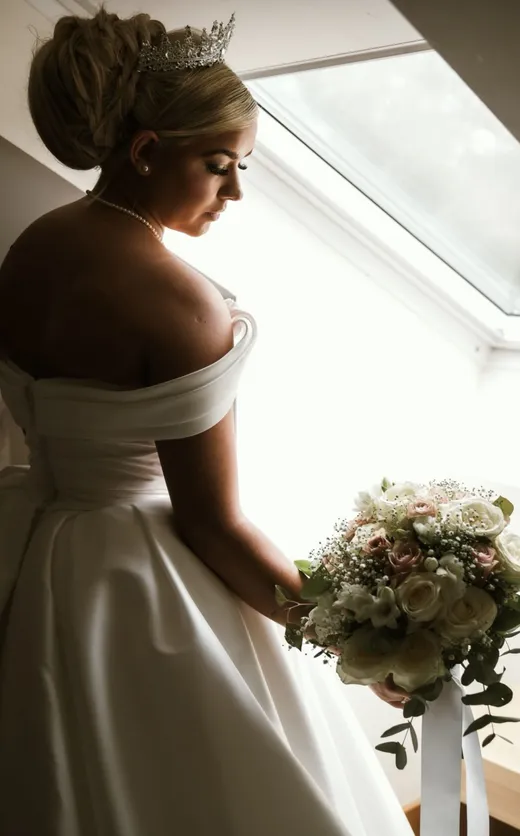 A stunning back view of a bride wearing an off-the-shoulder satin gown and a crystal tiara, holding a classic rose and baby's breath bouquet near a bright window.