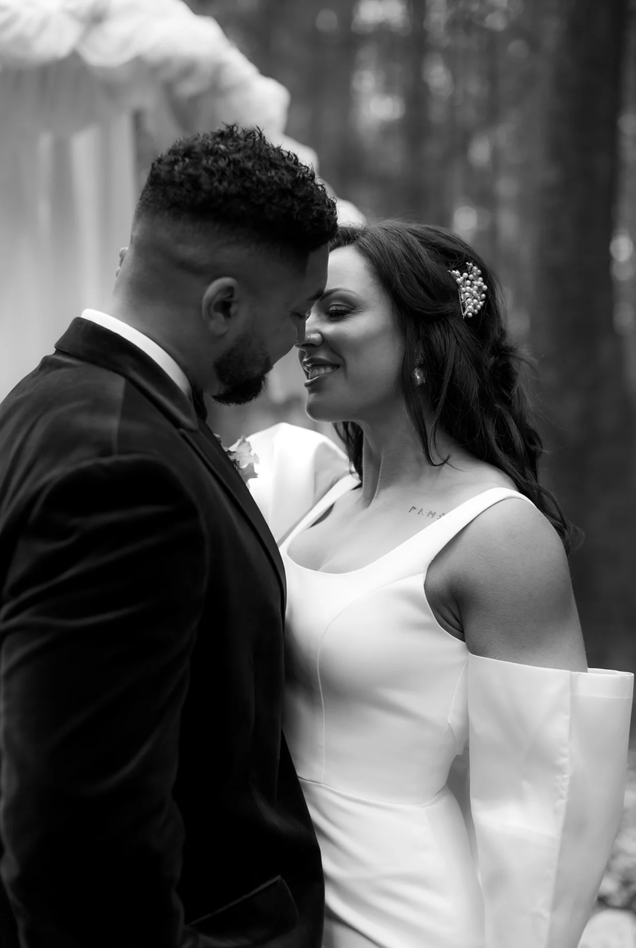 A newlywed couple shares an intimate moment with soft smiles, surrounded by a natural woodland setting in this timeless black and white wedding portrait.