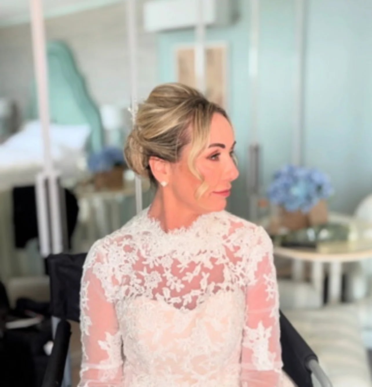 A profile portrait of a smiling bride getting ready in a bright room, wearing an intricate lace high-neck gown with her blonde hair styled in an elegant updo with face-framing tendrils.