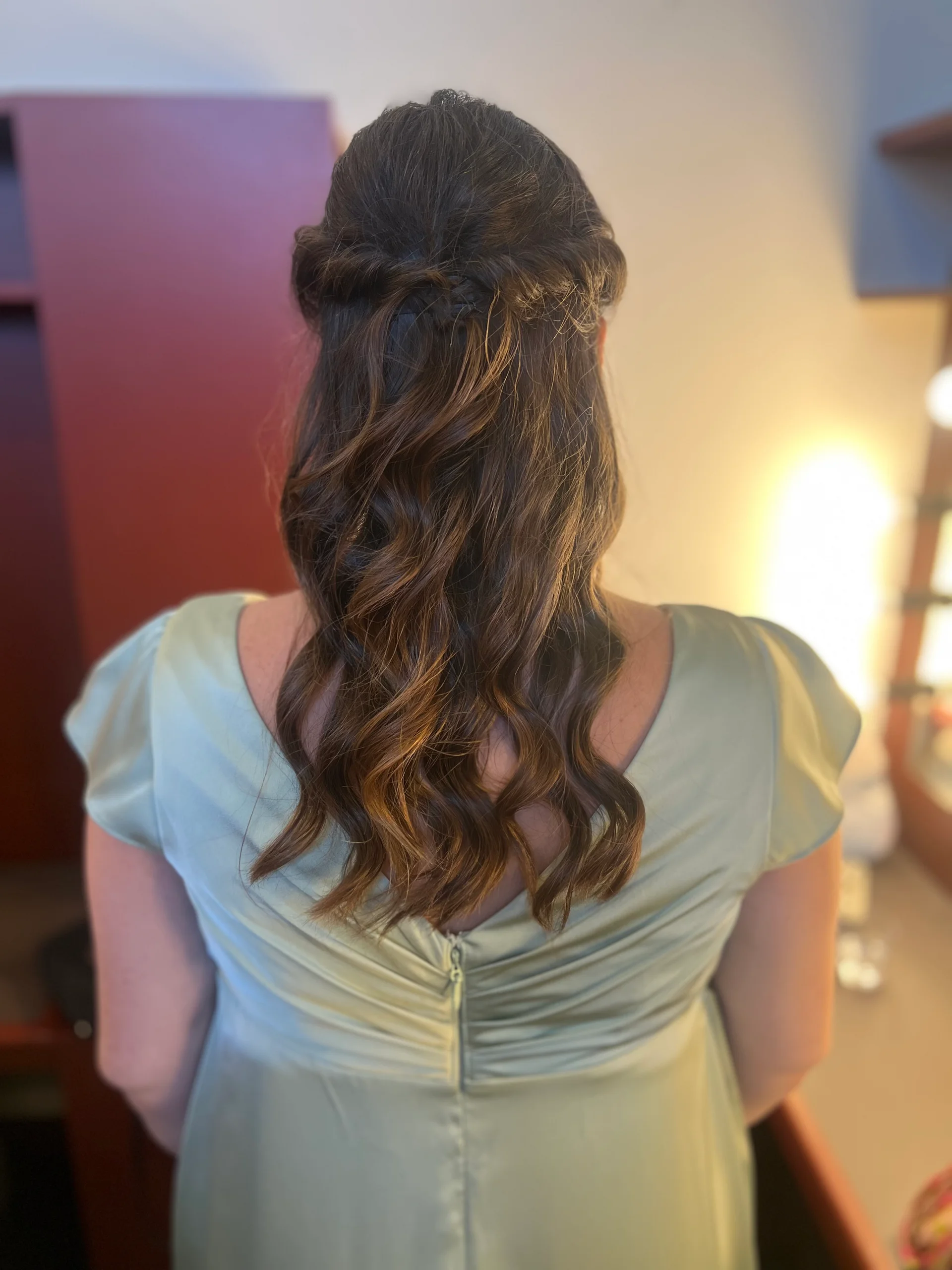 A back view of a bridesmaid wearing a sage green satin dress with her dark hair styled in a half-up half-down look with soft waves.