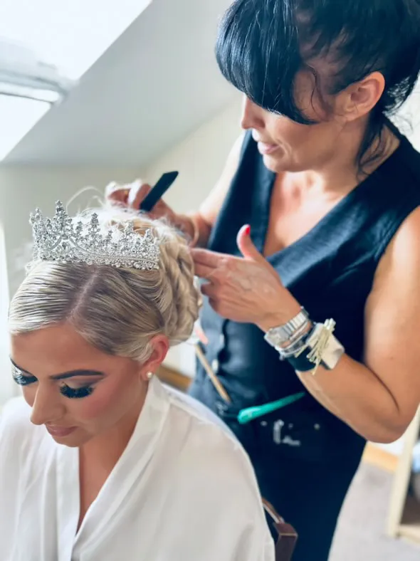 A bridal stylist carefully adjusts an intricate crystal tiara into a blonde bride's elegant low bun during morning preparations.