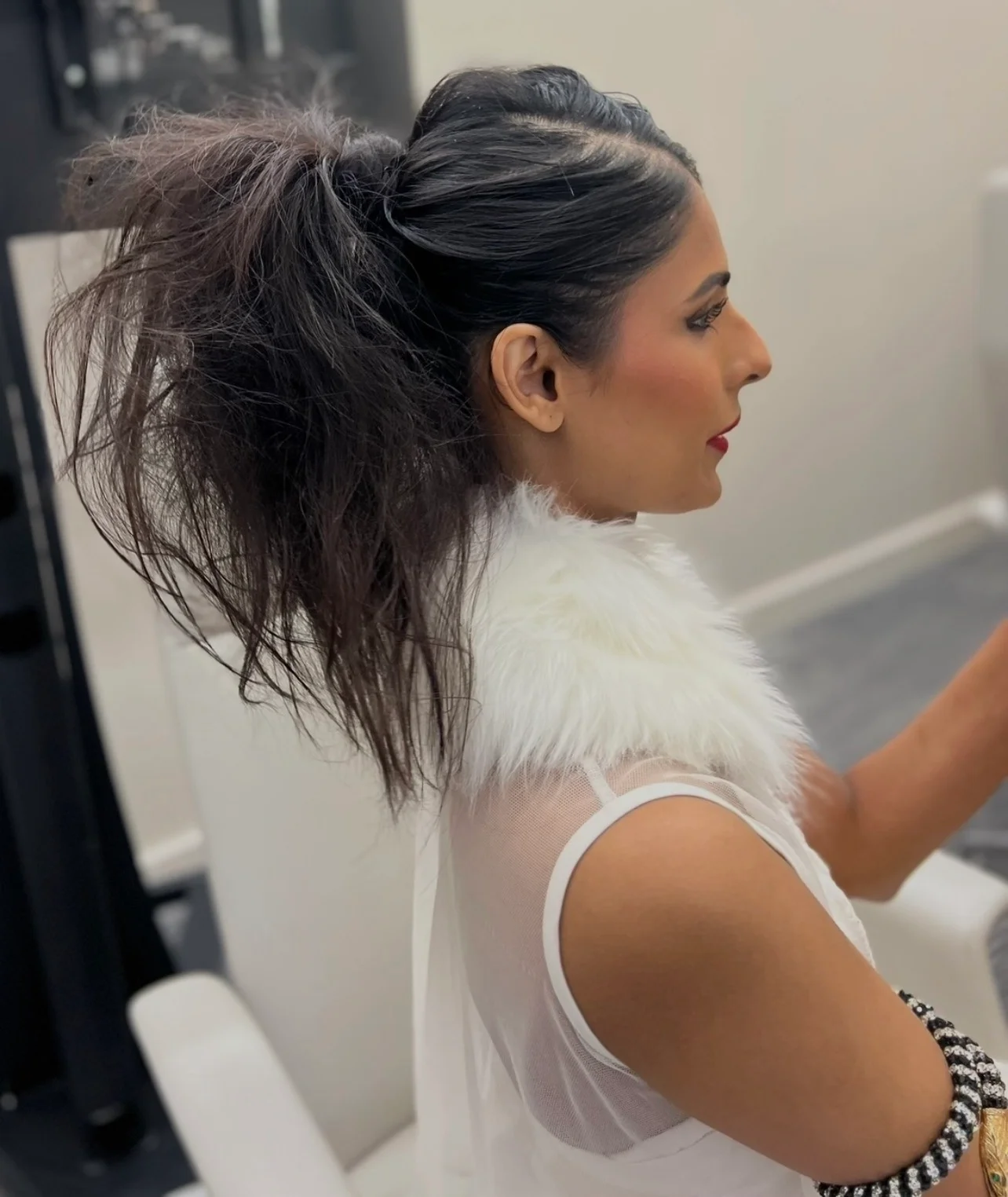 A side profile of a bride during morning preparations, showcasing her voluminous high ponytail hairstyle, soft glam makeup with a bold red lip, and a white top with a faux fur collar.