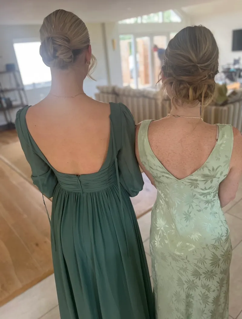Two bridesmaids showcase their elegant hairstyles and matching green dresses from behind while getting ready indoors.