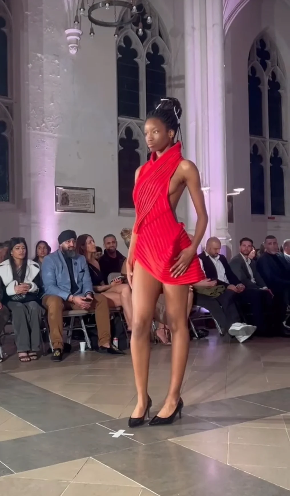 A model walks the runway in a striking red high-necked, draped minidress with a high thigh slit inside a grand church venue.