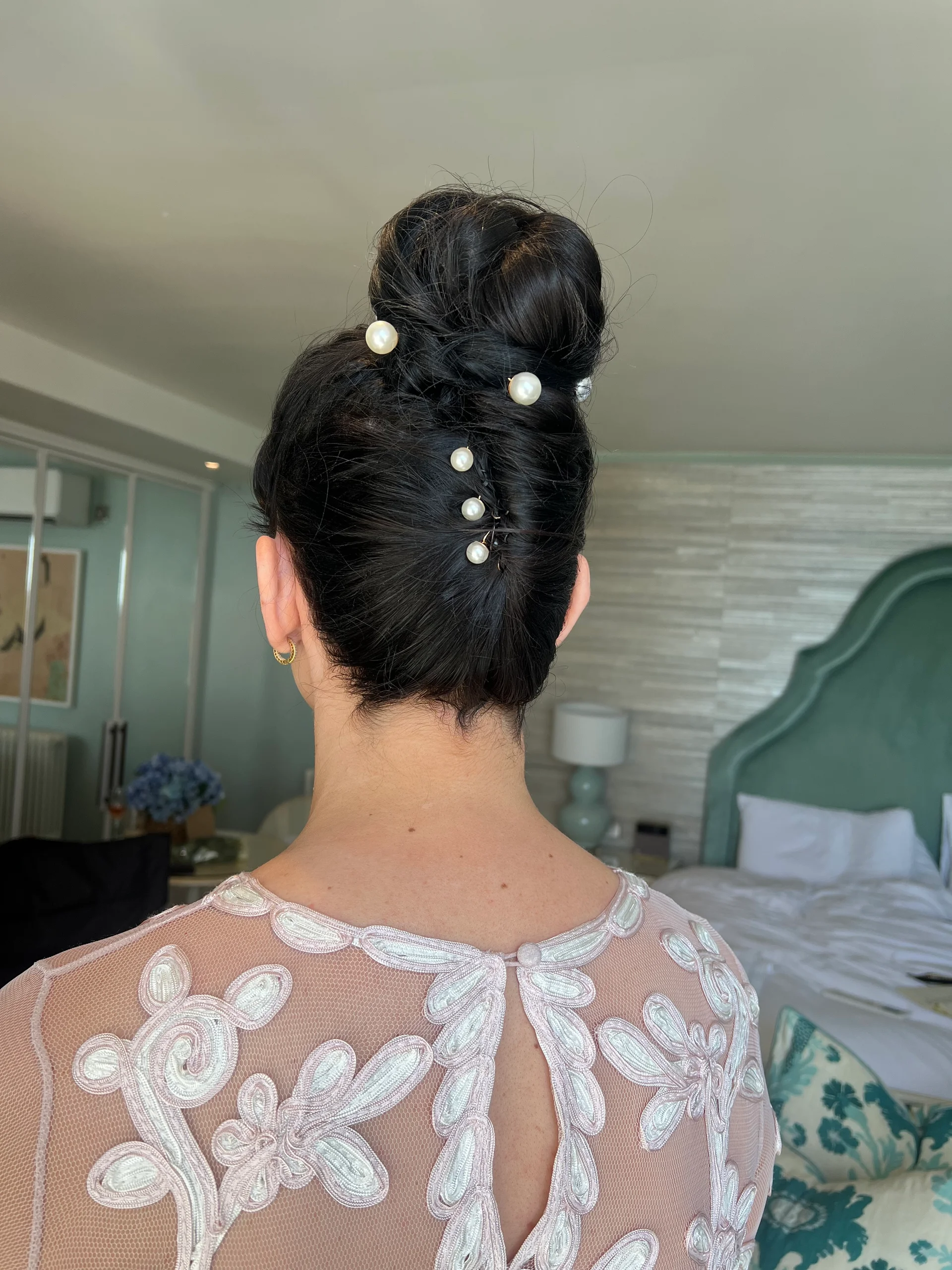 A back view of a bride showcasing an intricate high bun adorned with pearl hair pins, paired with a sheer pink dress featuring white floral appliqué details.