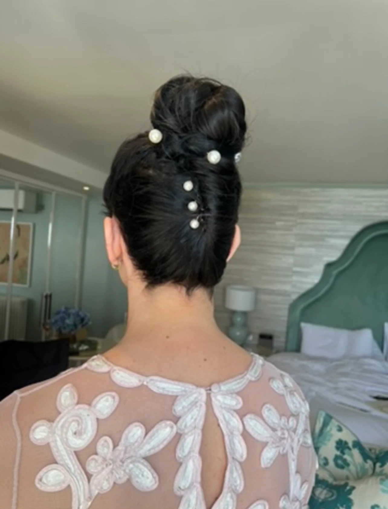 A back view of a bride showcasing an intricate high bun hairstyle adorned with delicate pearl hair pins, paired with a sheer dress featuring floral applique details.