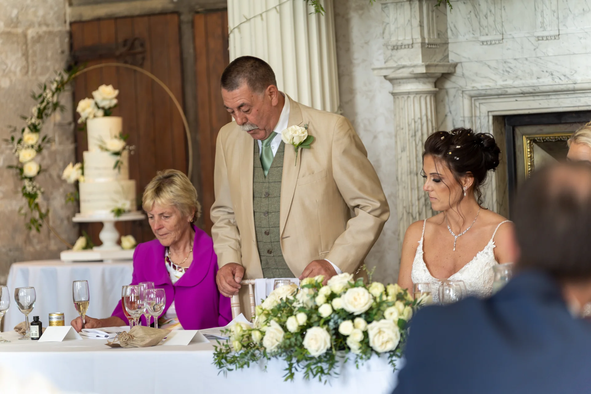 The father of the bride stands to deliver his speech at the top table, flanked by the mother of the bride and the bride during the wedding reception.