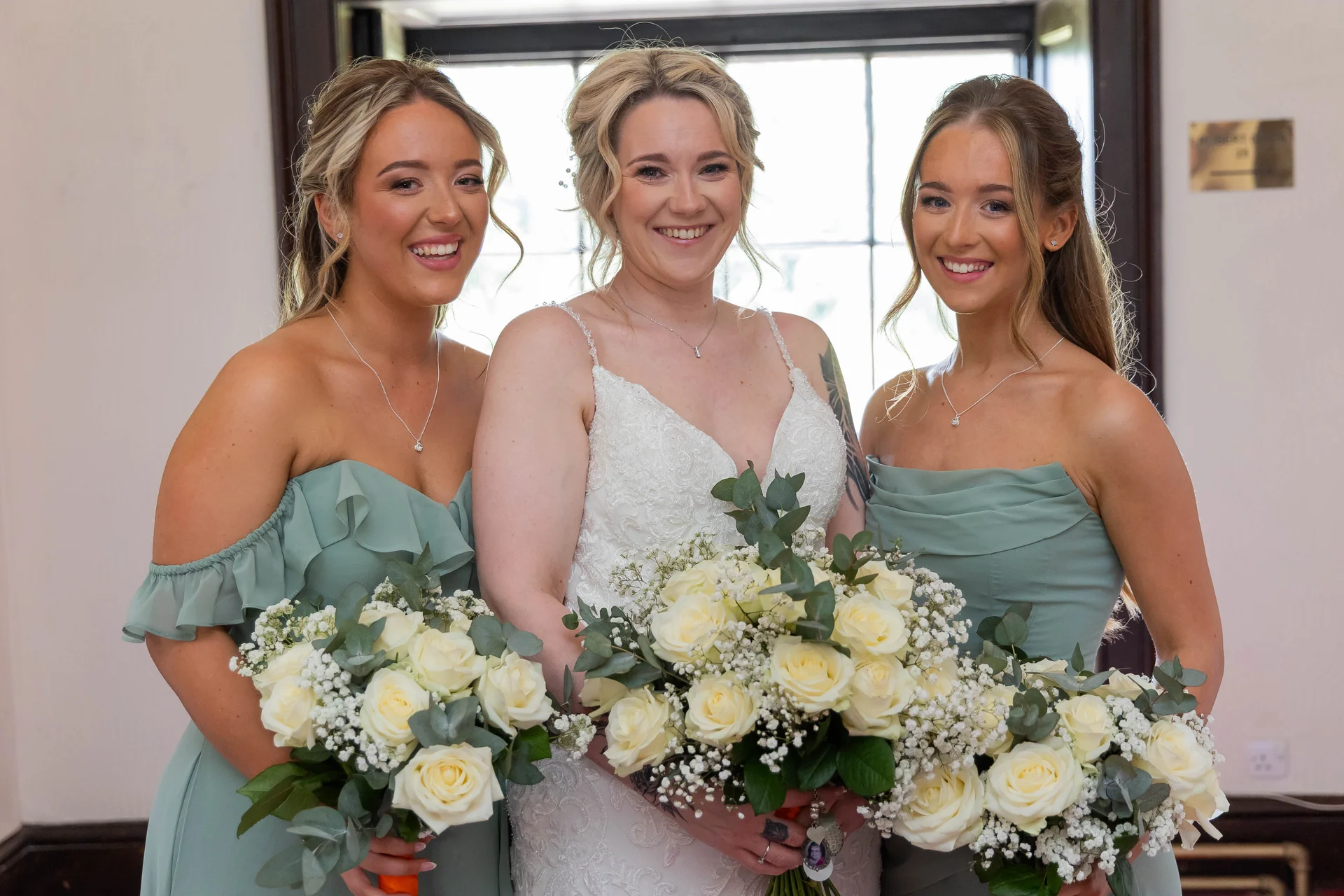 A smiling bride wearing a lace gown and two bridesmaids in matching sage dresses holding white rose bouquets pose indoors near a window.
