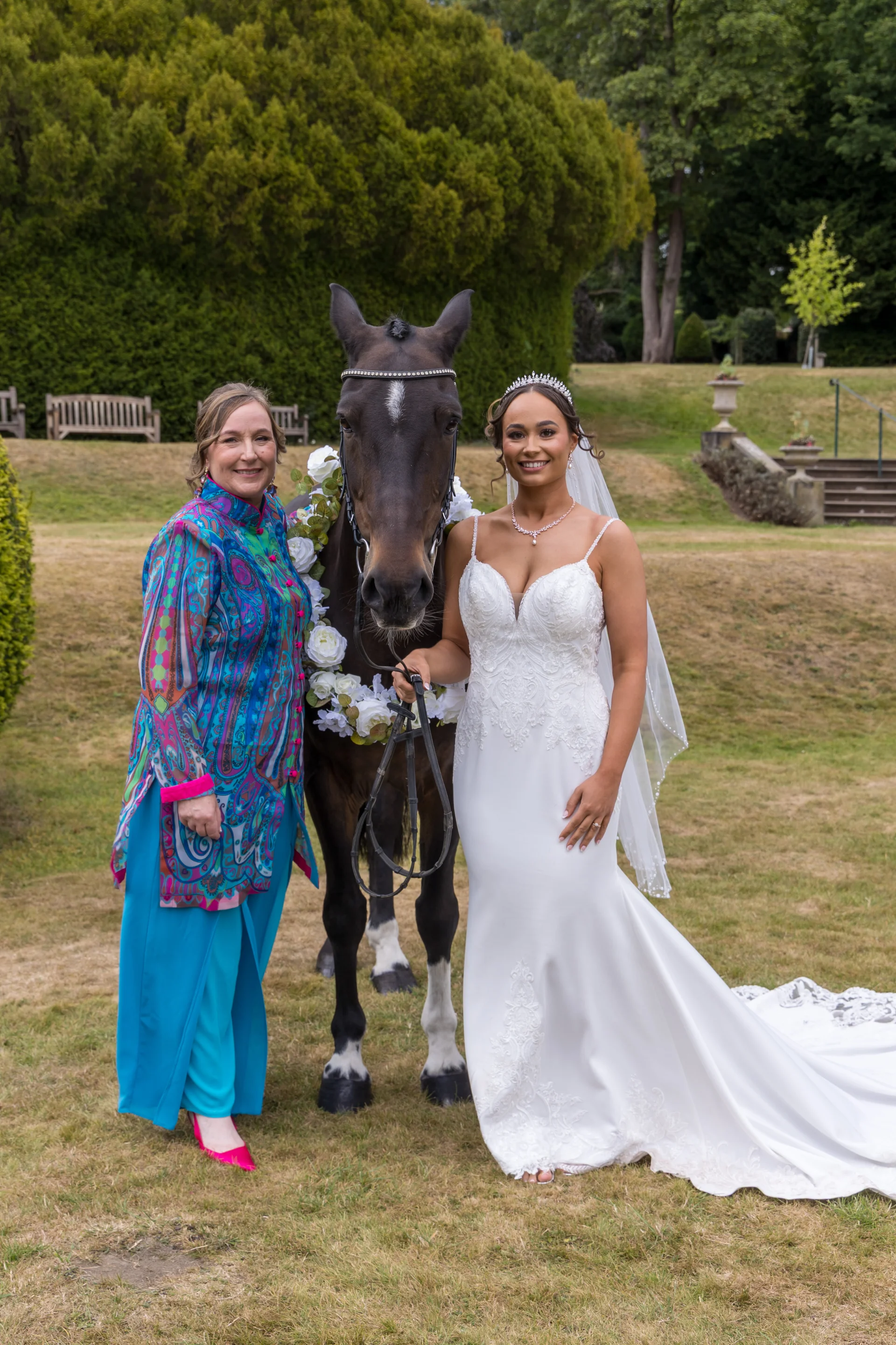 A smiling bride in a white lace fit-and-flare gown stands alongside a guest and a horse adorned with white floral garlands in a scenic garden.