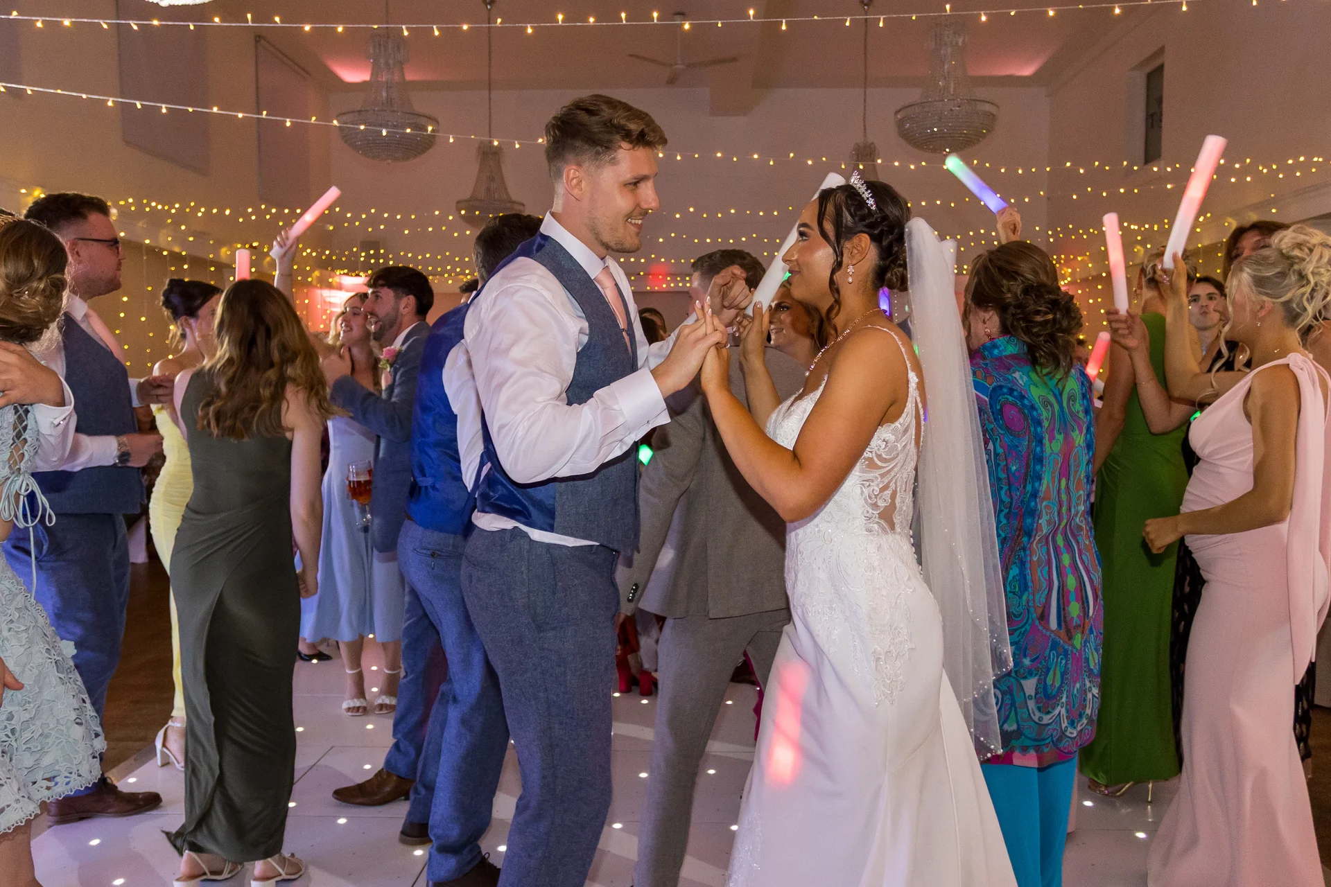 The bride and groom share a joyful first dance on a lit dance floor surrounded by cheering guests waving glow sticks under a fairy light canopy.
