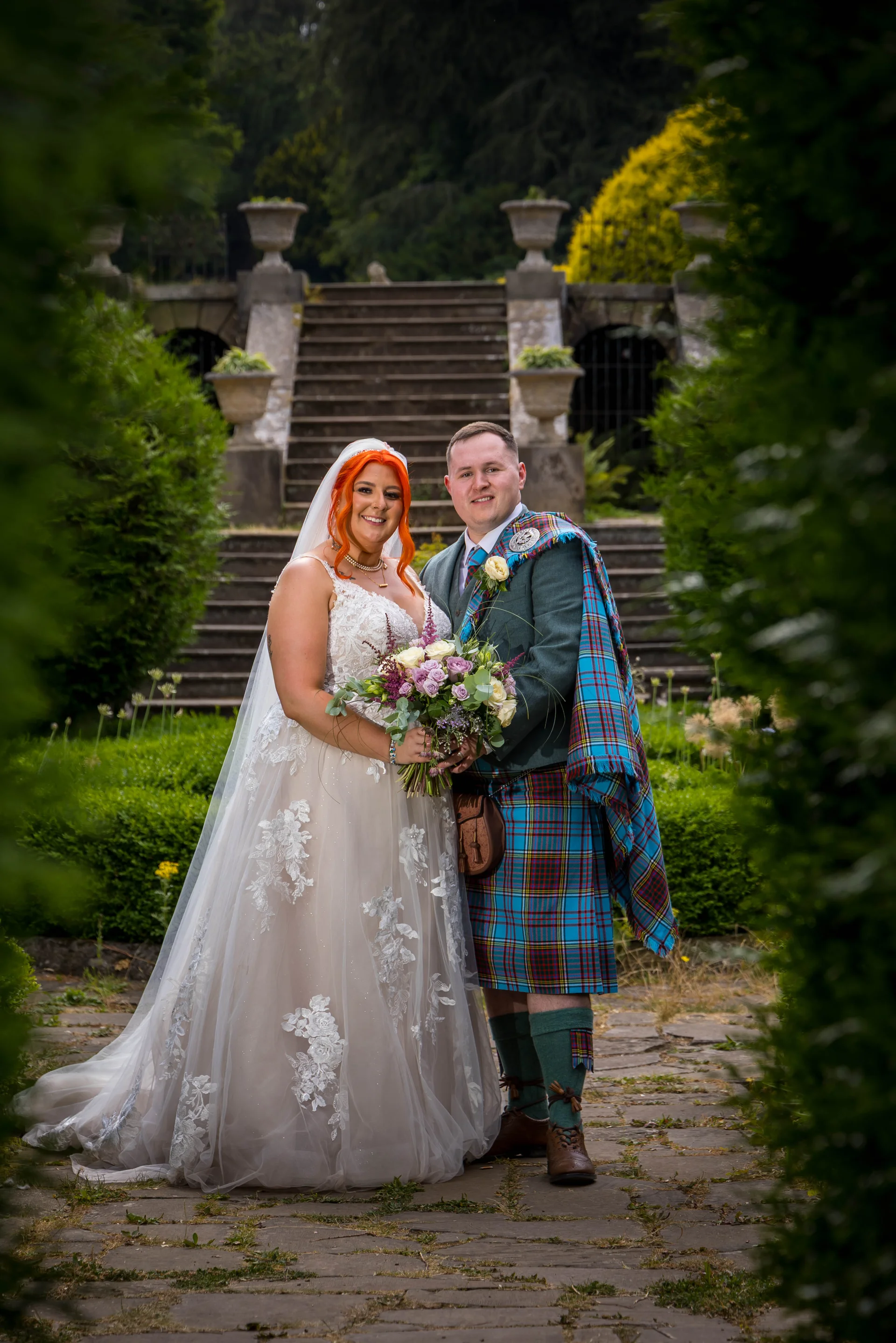 A newlywed couple poses in a manicured garden framing a stone staircase, with the bride wearing a lace-detailed a-line gown and vibrant red hair, and the groom in a traditional tartan kilt and tweed jacket.