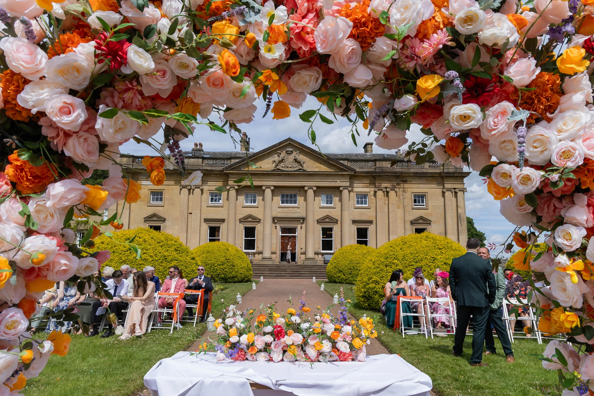 An outdoor wedding ceremony featuring a vibrant floral arch framing a grand stone country house and seated guests on the lawn.