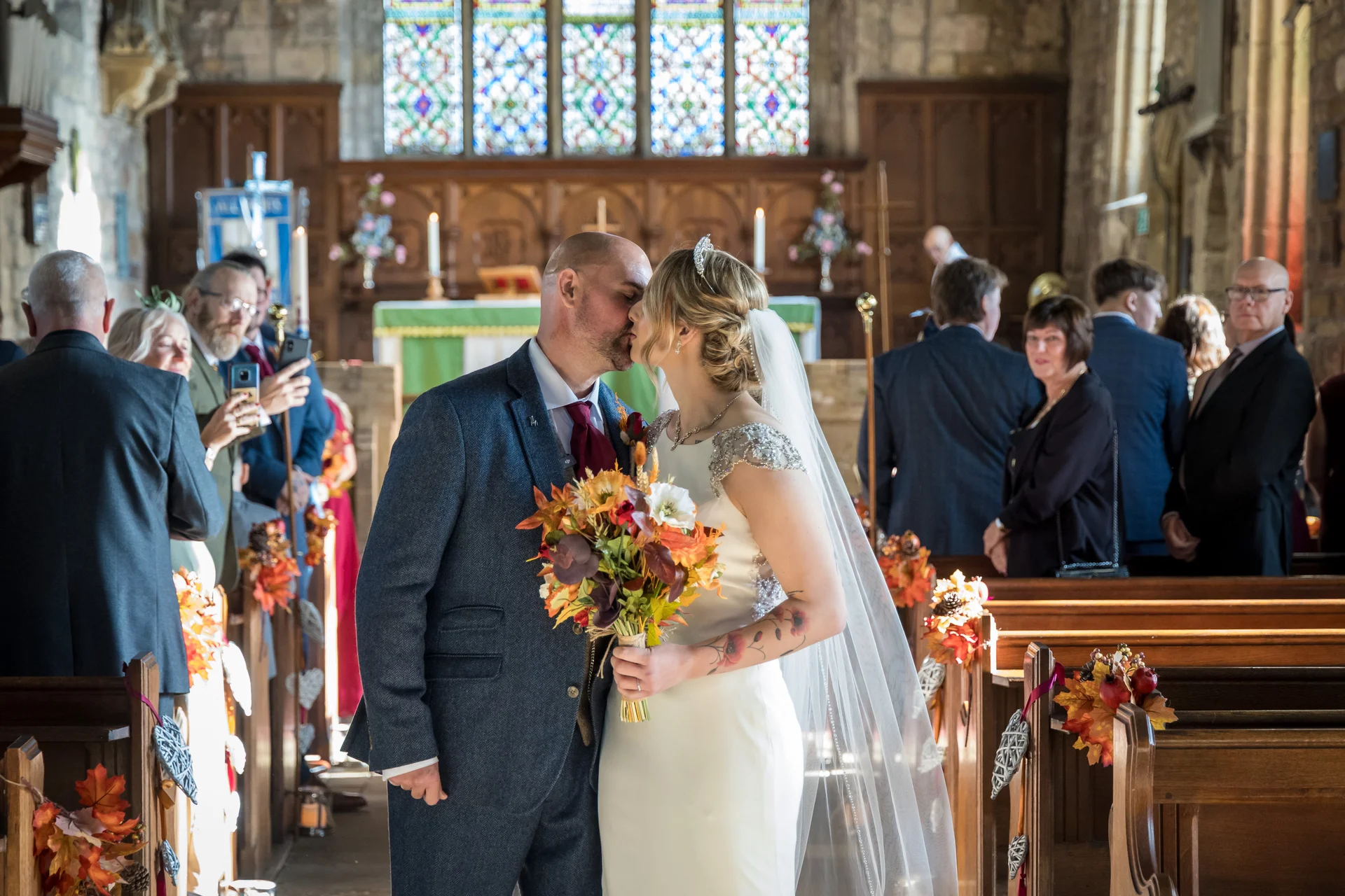 A newlywed couple share their first kiss at the altar of a traditional church, surrounded by family, wooden pews, and vibrant autumn floral decorations.